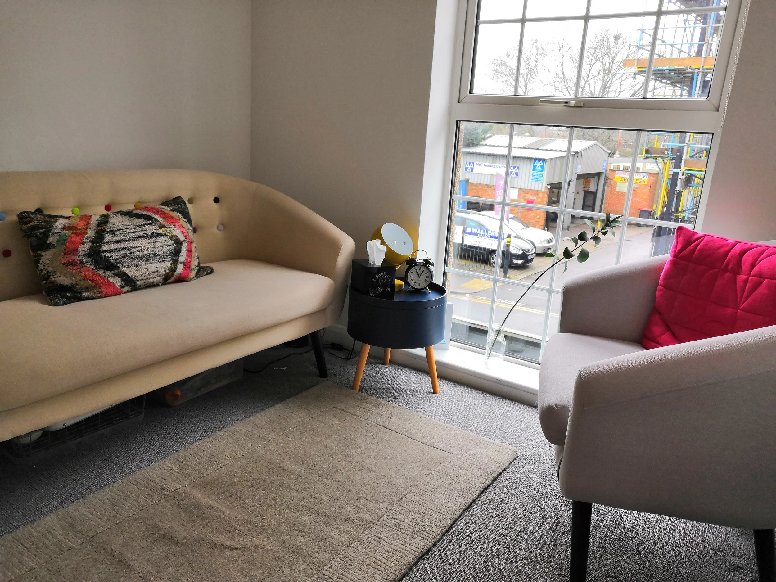 A cozy living room with a beige couch with a colorful cushion and a pink blanket on a gray armchair, a small black side table with a clock, tissue box, and mirror, a large window showing a street scene with cars and a construction site outside.