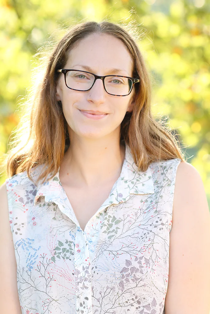 Portrait of a young woman with light brown hair, glasses, and a sleeveless patterned blouse, smiling outdoors with blurred green foliage in the background.
