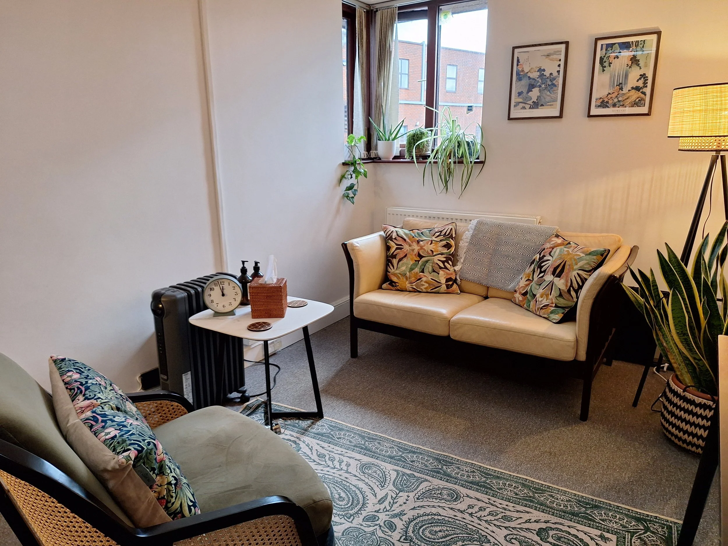 Cozy living room with a cream sofa and patterned cushions, a rattan armchair with floral pillow, a small white table with a clock and hand sanitizer, large window with plants on the sill, framed artwork on the wall, and a floor lamp nearby.