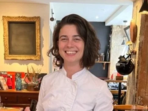 Une femme souriante avec des cheveux bruns bouclés, portant une blouse blanche, dans un intérieur décoré de façon éclectique.