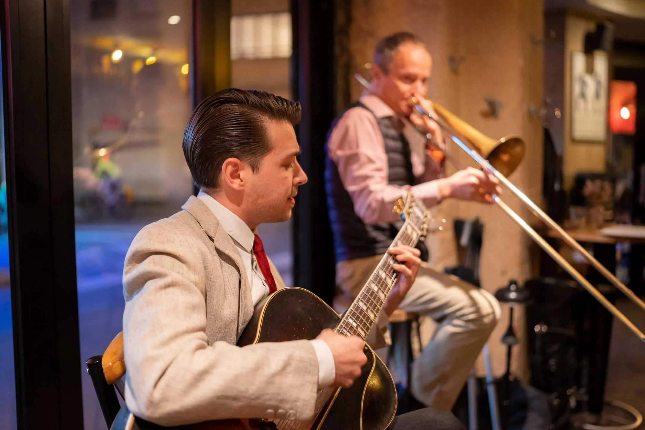 Deux musiciens d'inspiration jazz jouent dans un café, l'un avec une guitare acoustique et l'autre avec un trombone.