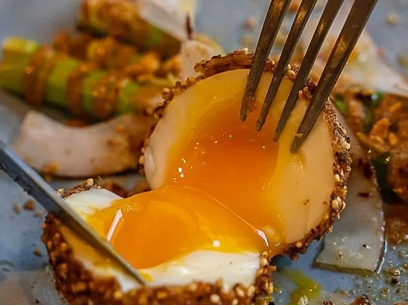 Oeuf mollet avec le jaune coulant, entouré de graines ou d'épices, avec des légumes sautés en arrière-plan.