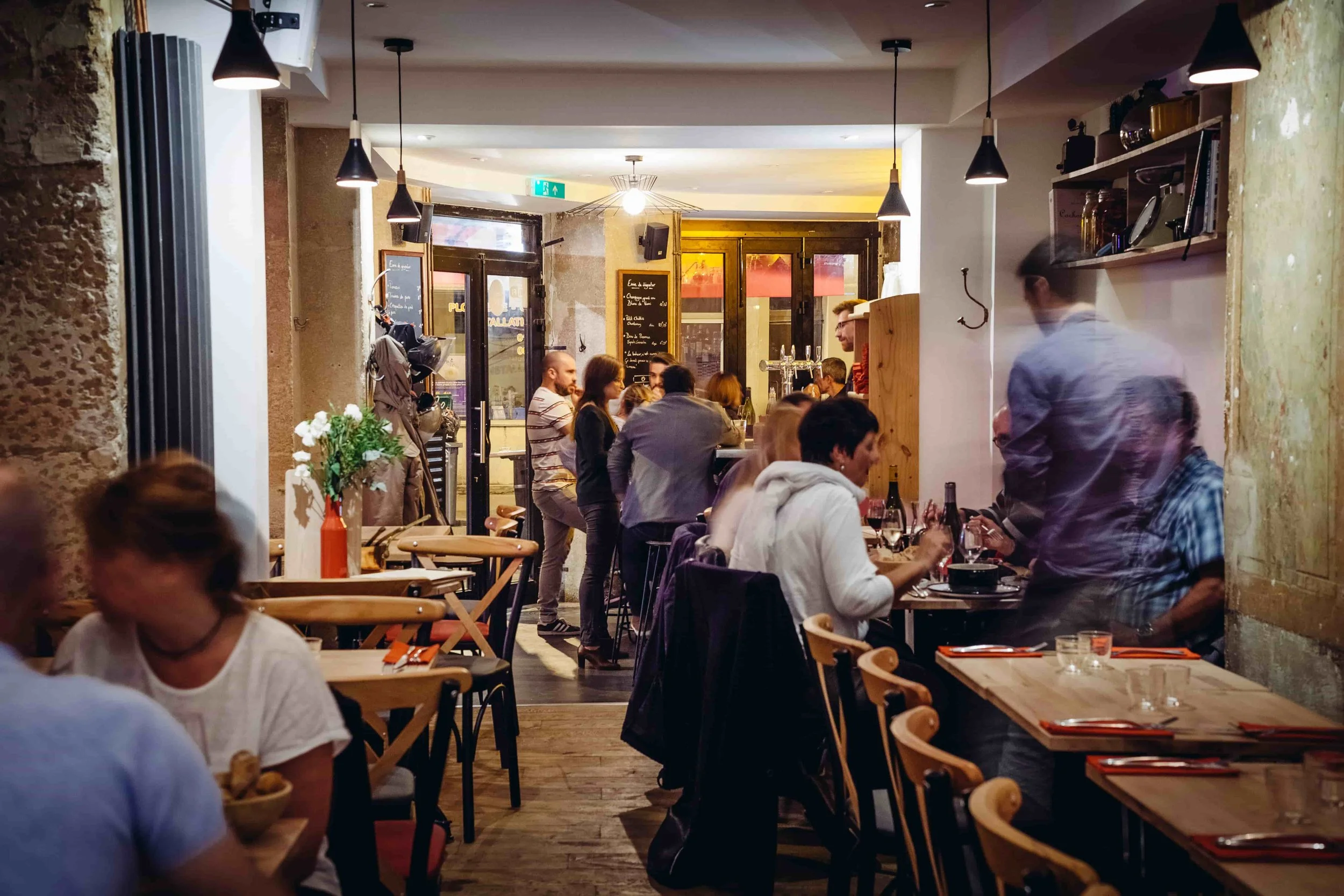 Intérieur d'un restaurant animé avec des clients discutant et mangeant, décor en pierre et bois, éclairage suspendu chaud.