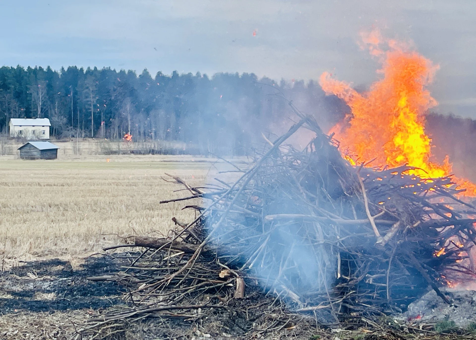 Pääsiäiskokko palaa ja savuaa. Taustalla peltomaisema, metsää ja puurakennus