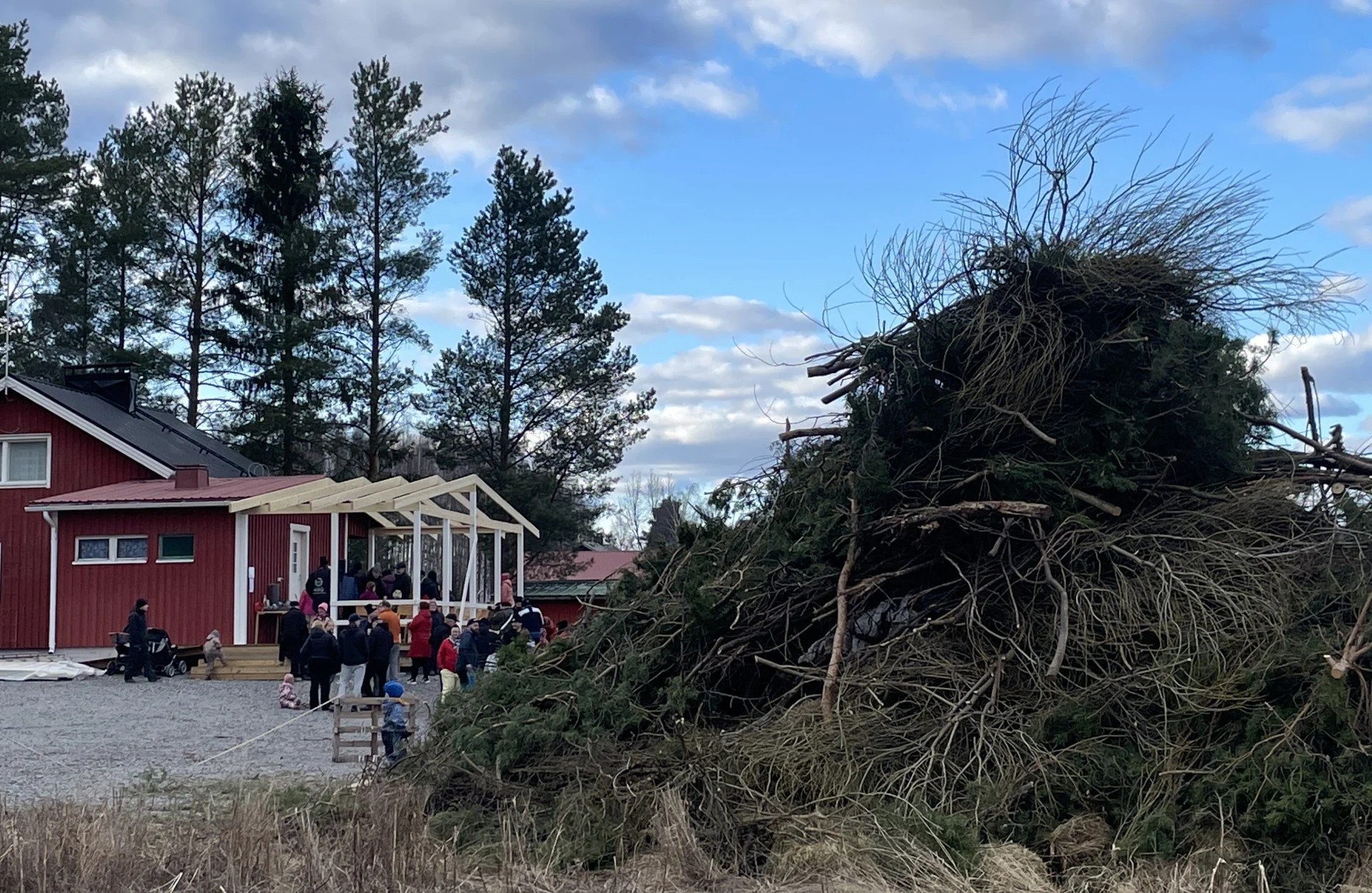 Kuva pääsiäiskokosta joka ei vielä ole sytytetty, taustalla ihmisjoukko ja punainen puinen kylätalo ja sininen taivas