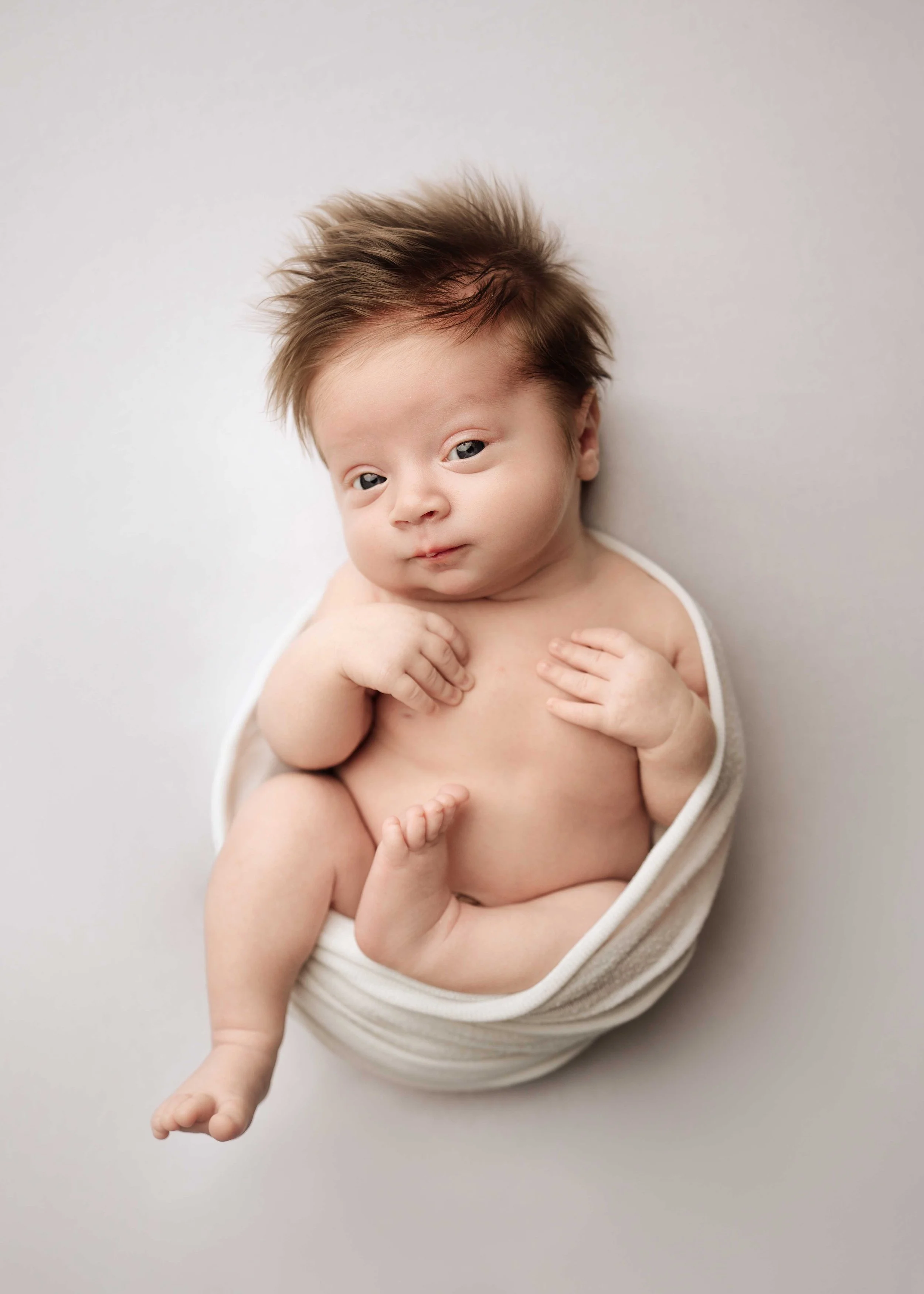 awake newborn baby boy lies in a white wrap that goes around his body