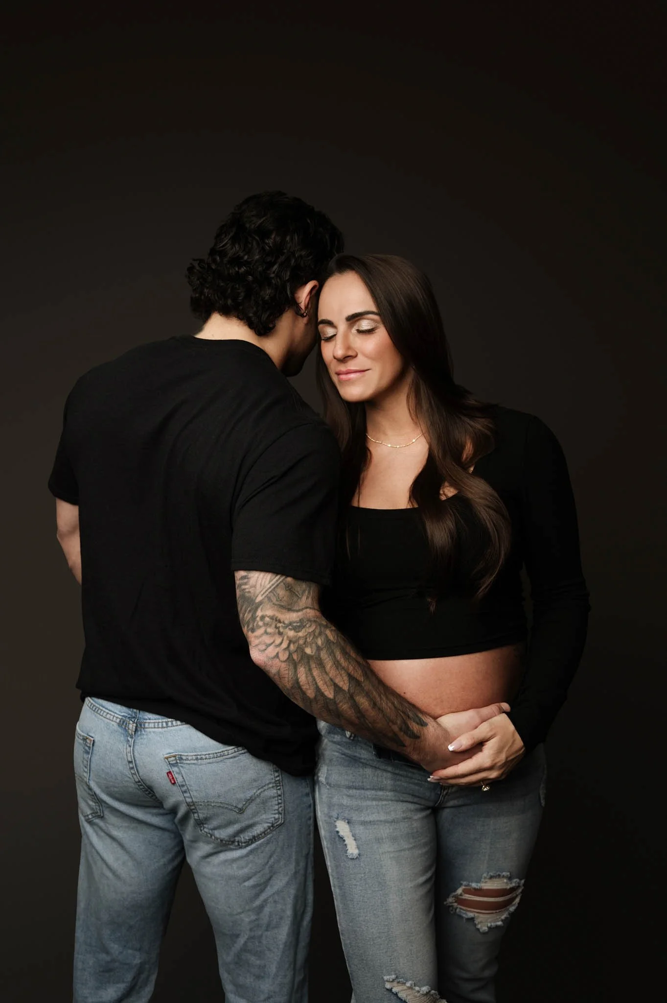 woman and partner, both wearing black shirts and jeans, pose in a Fairfield county, ct maternity session against a gray backdrop