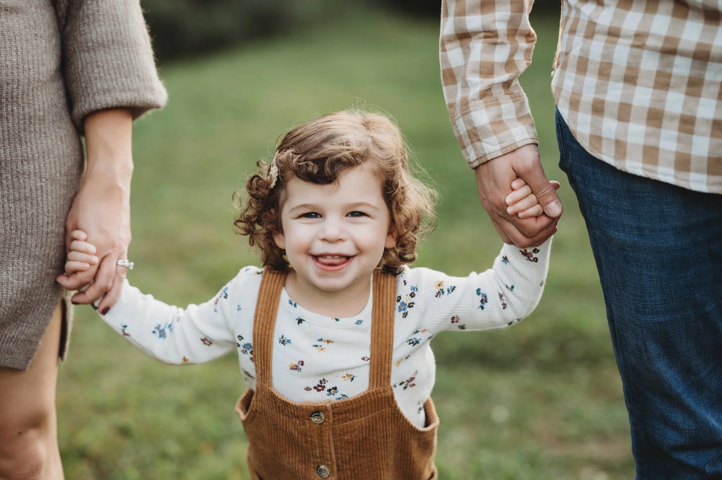 little girl in corduroy jumper and floral shirt holds both parents hands and smiles in a ridgefield, ct park