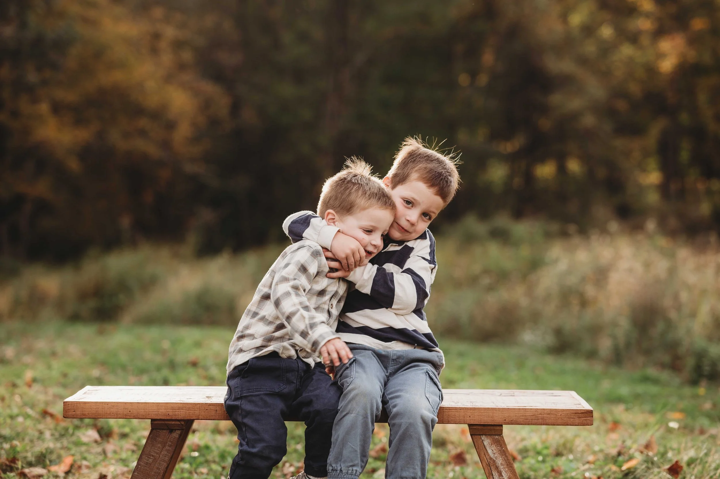 hugging brothers sit on a bench surrounded by a fall backdrop, hugging