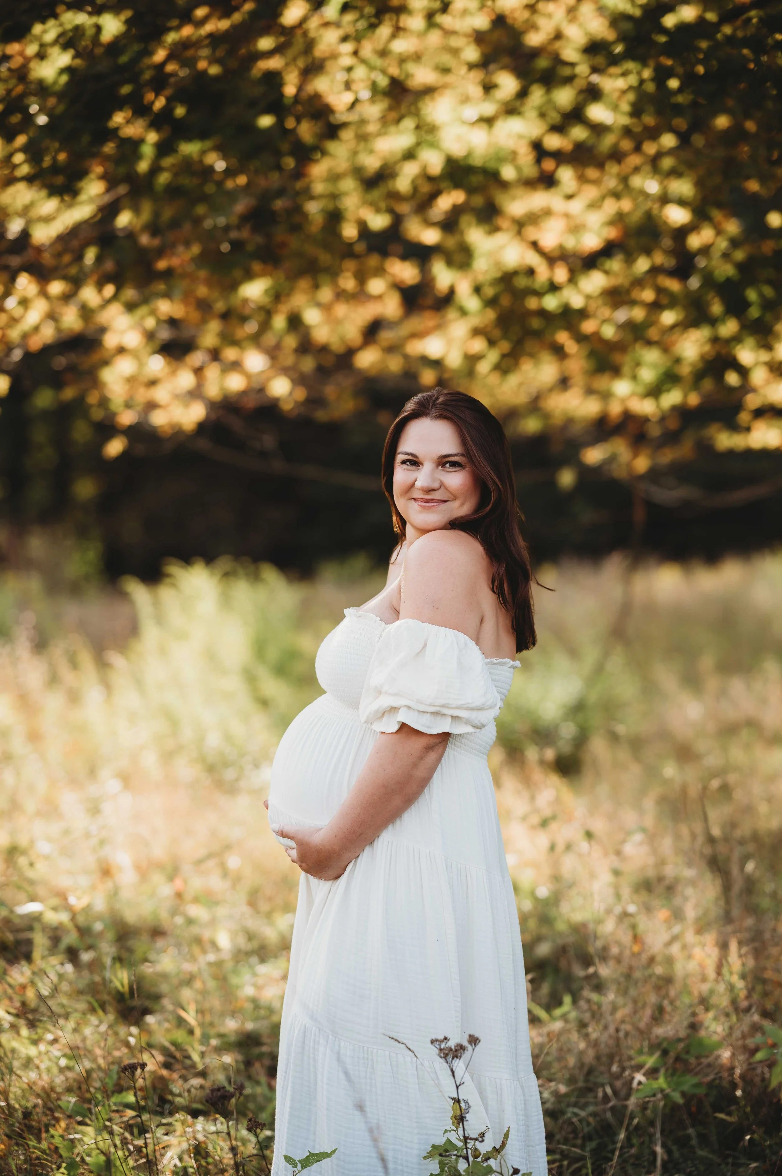 Pregnant woman in white dress holds belly and looks at the camera in a Fairfield County, ct field