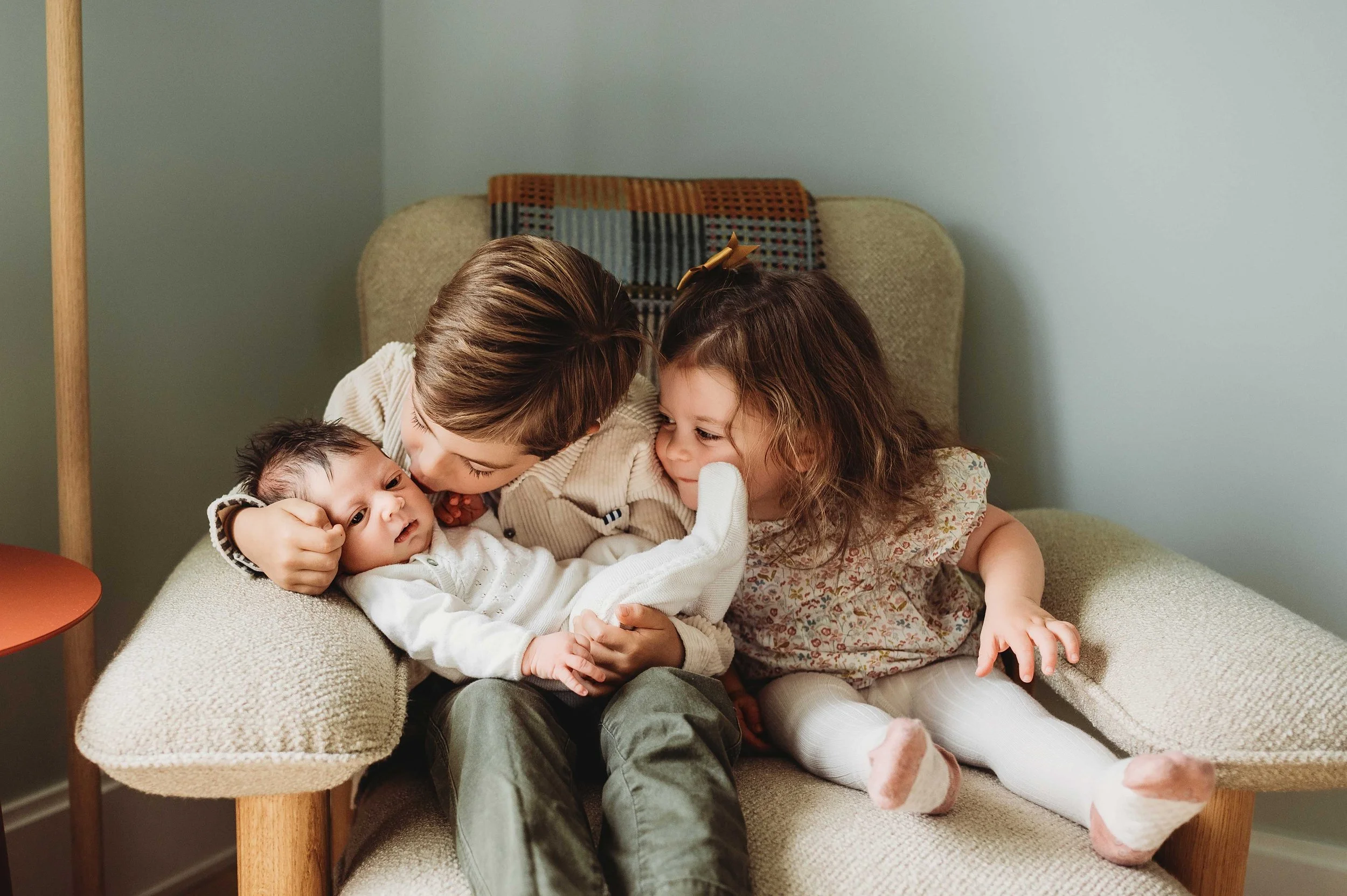 two siblings hold newborn baby sister, brother leaning over to kiss the baby, in an in-home lifestyle photoshoot in greenwich, ct