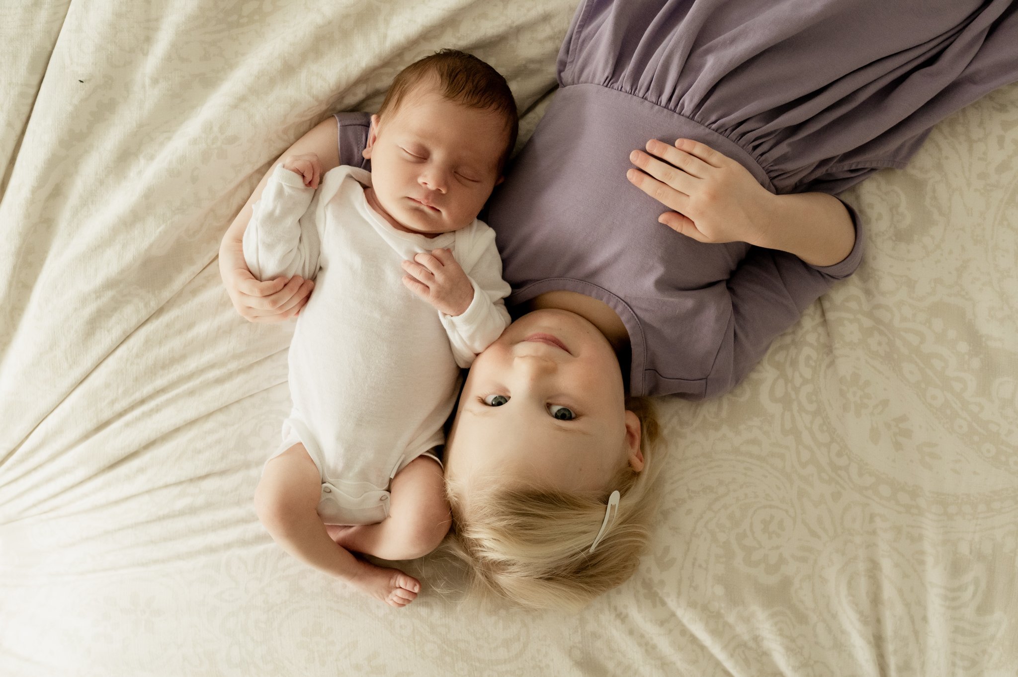 Toddler snuggles baby sister on bed