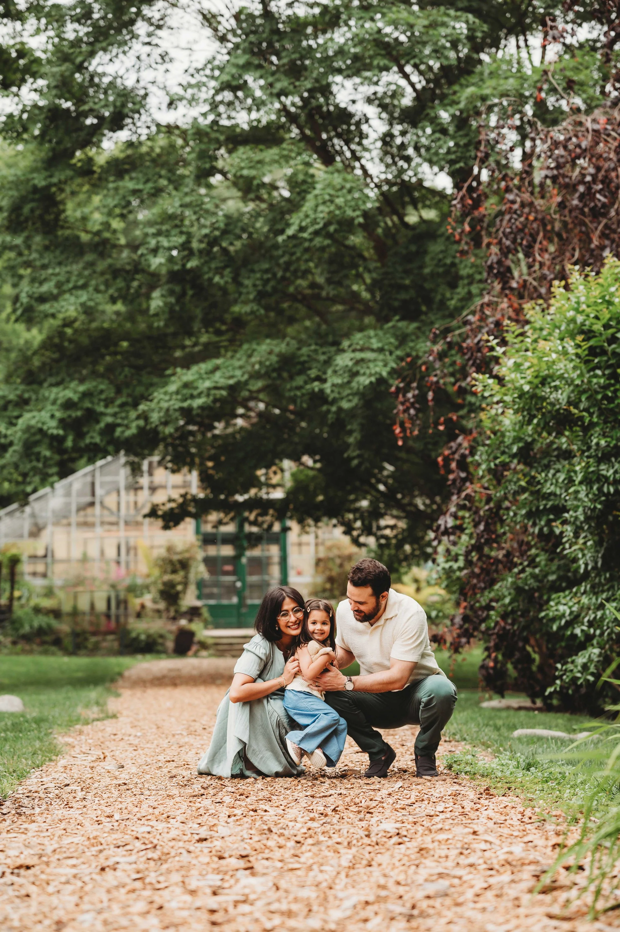 family of three crouches down to play with young daughter in park, stamford, ct