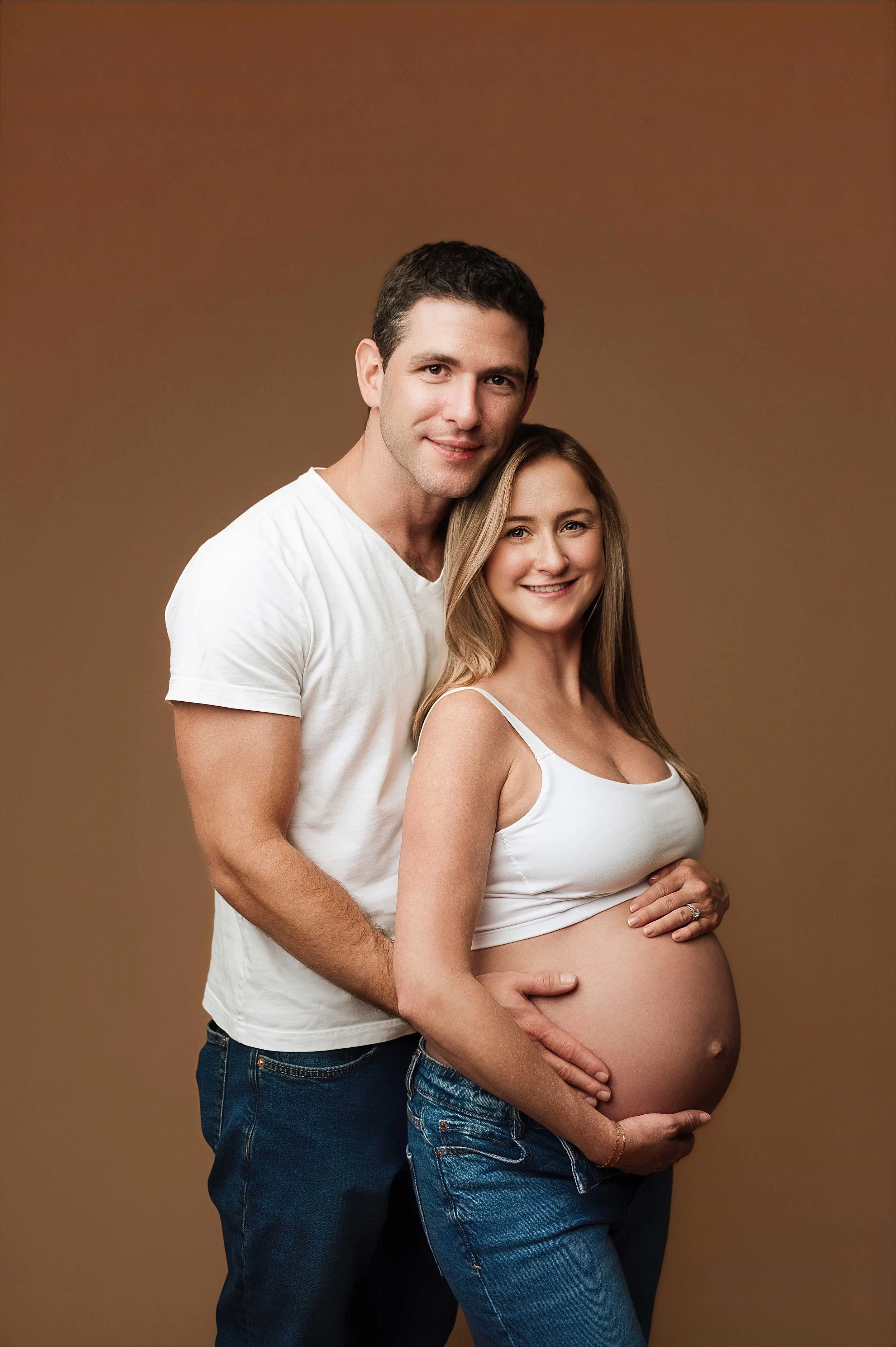 pregnant woman wearing crop tank and jeans poses with husband in front of brown backdrop, ridgefield, ct photography studio