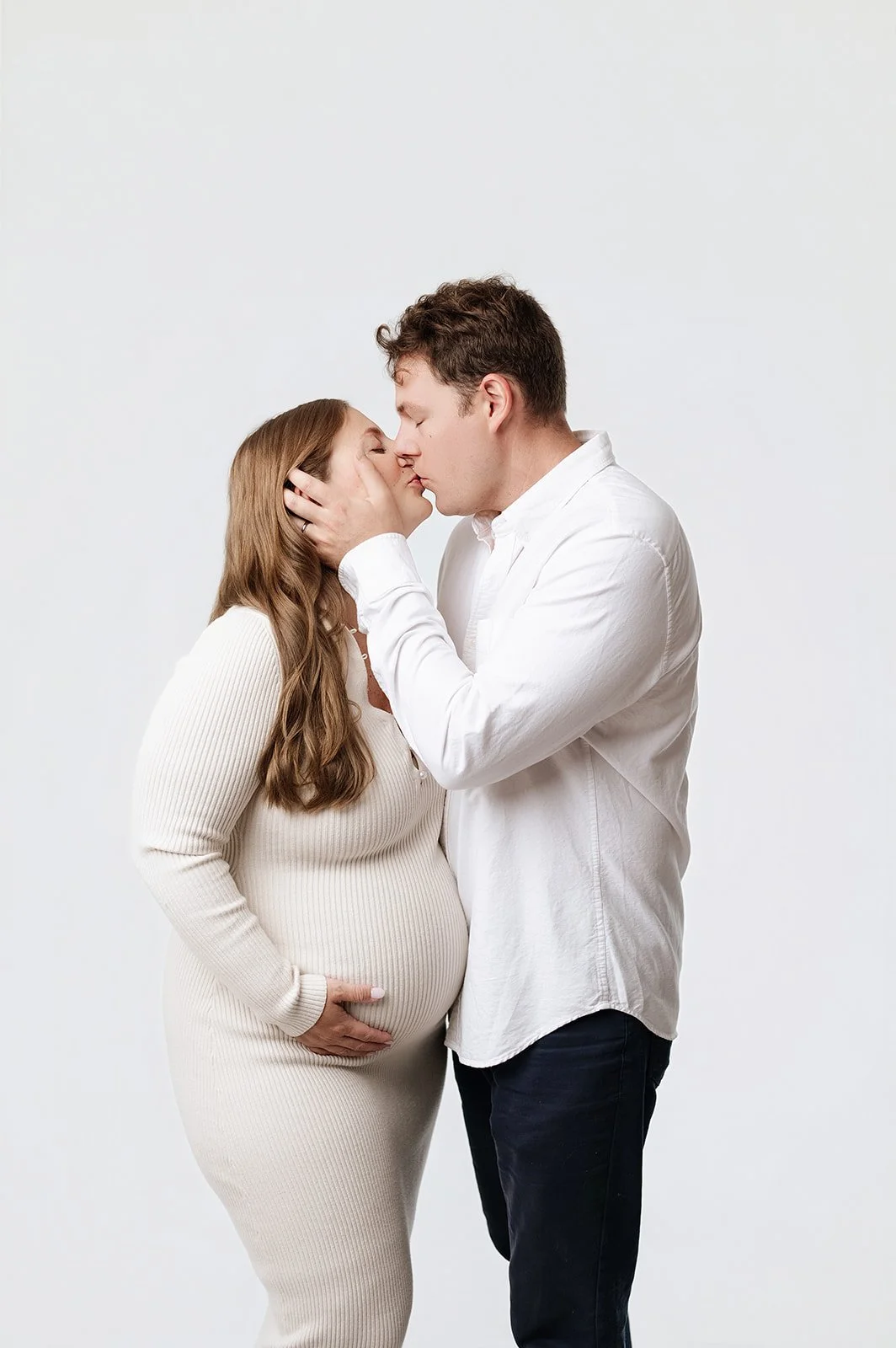 expecting couple in cream colors poses in front of white backdrop, ridgefield, ct studio