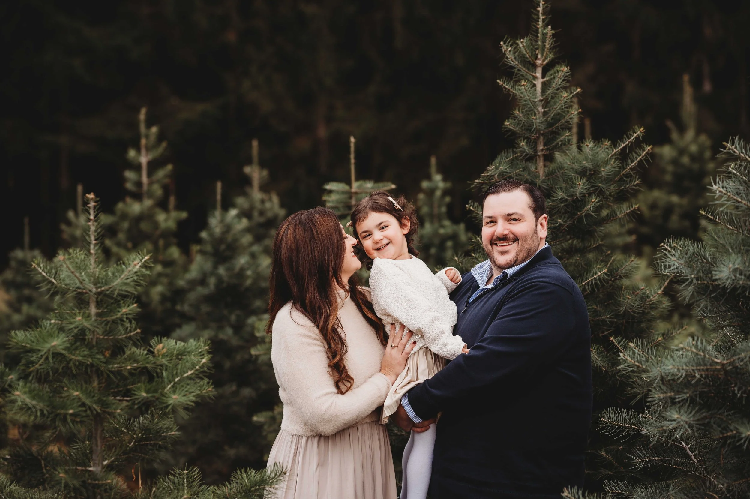 Mom, dad, and 3 year old girl playfully pose in front of christmas trees at a tree farm