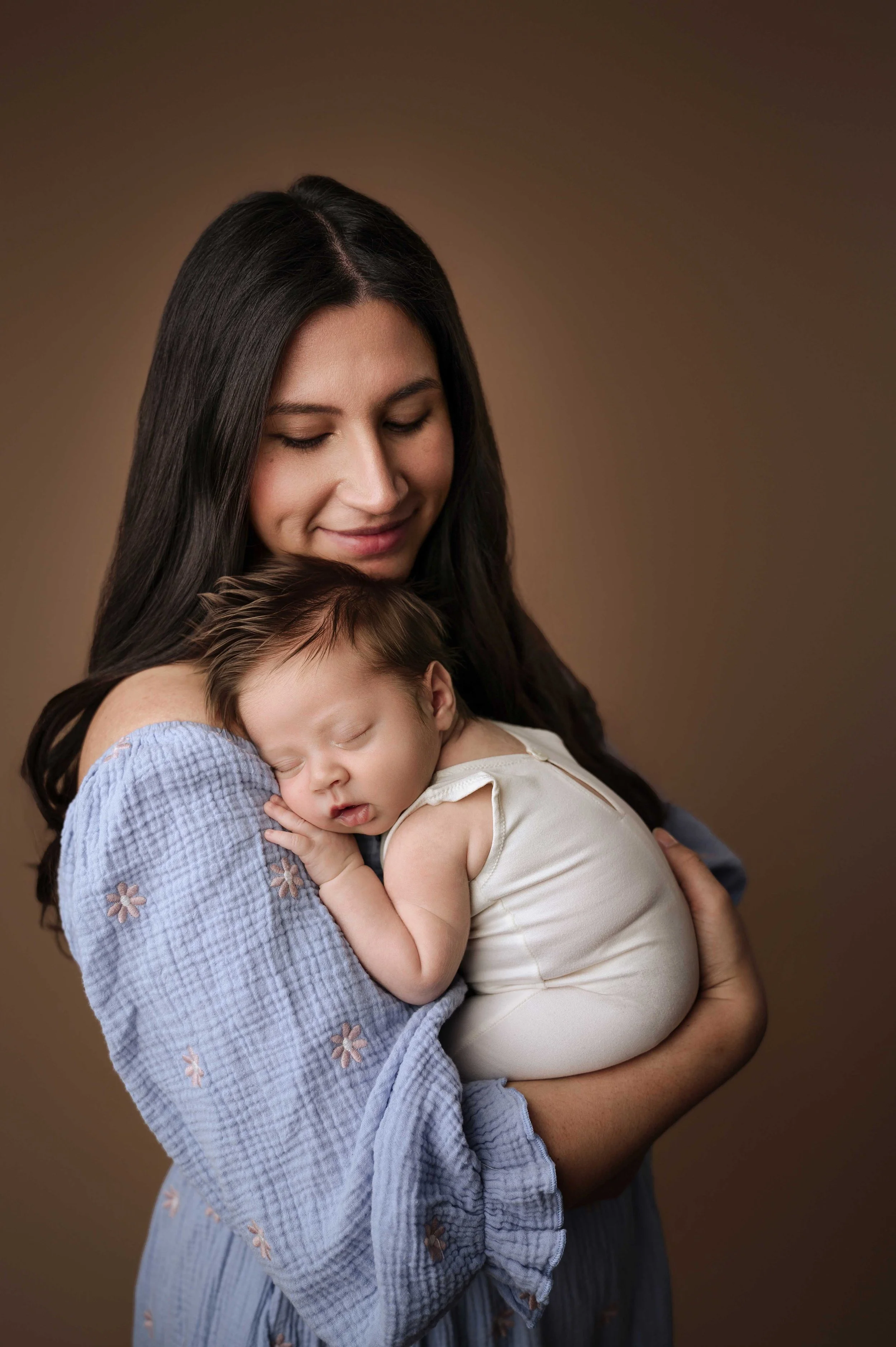 Mother holds baby against a brown backdrop. baby is curled up on her chest, against her shoulder.