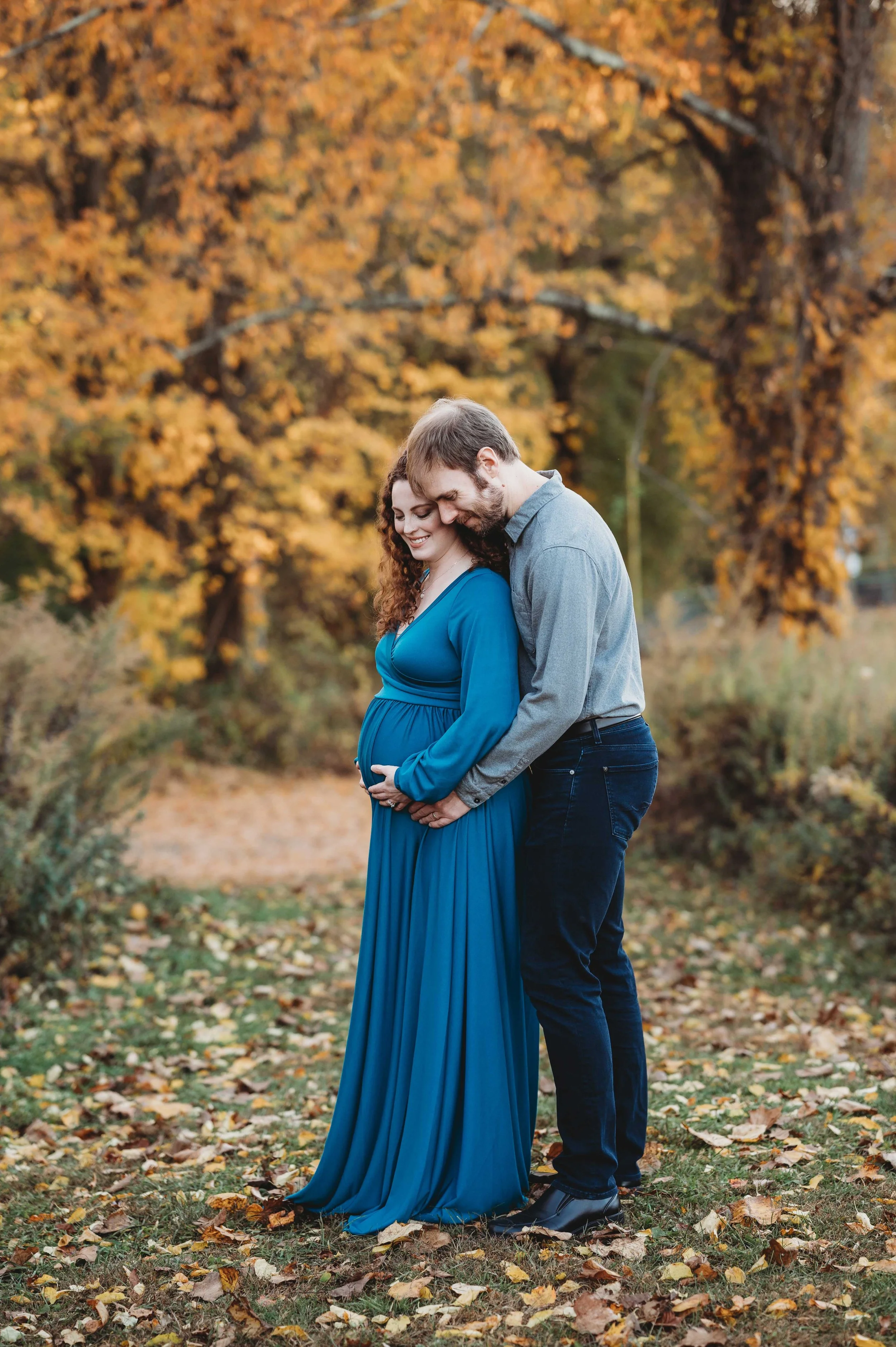 woman in blue dress poses with husband with fall foliage behind her in Ridgefield CT during her maternity photoshoot