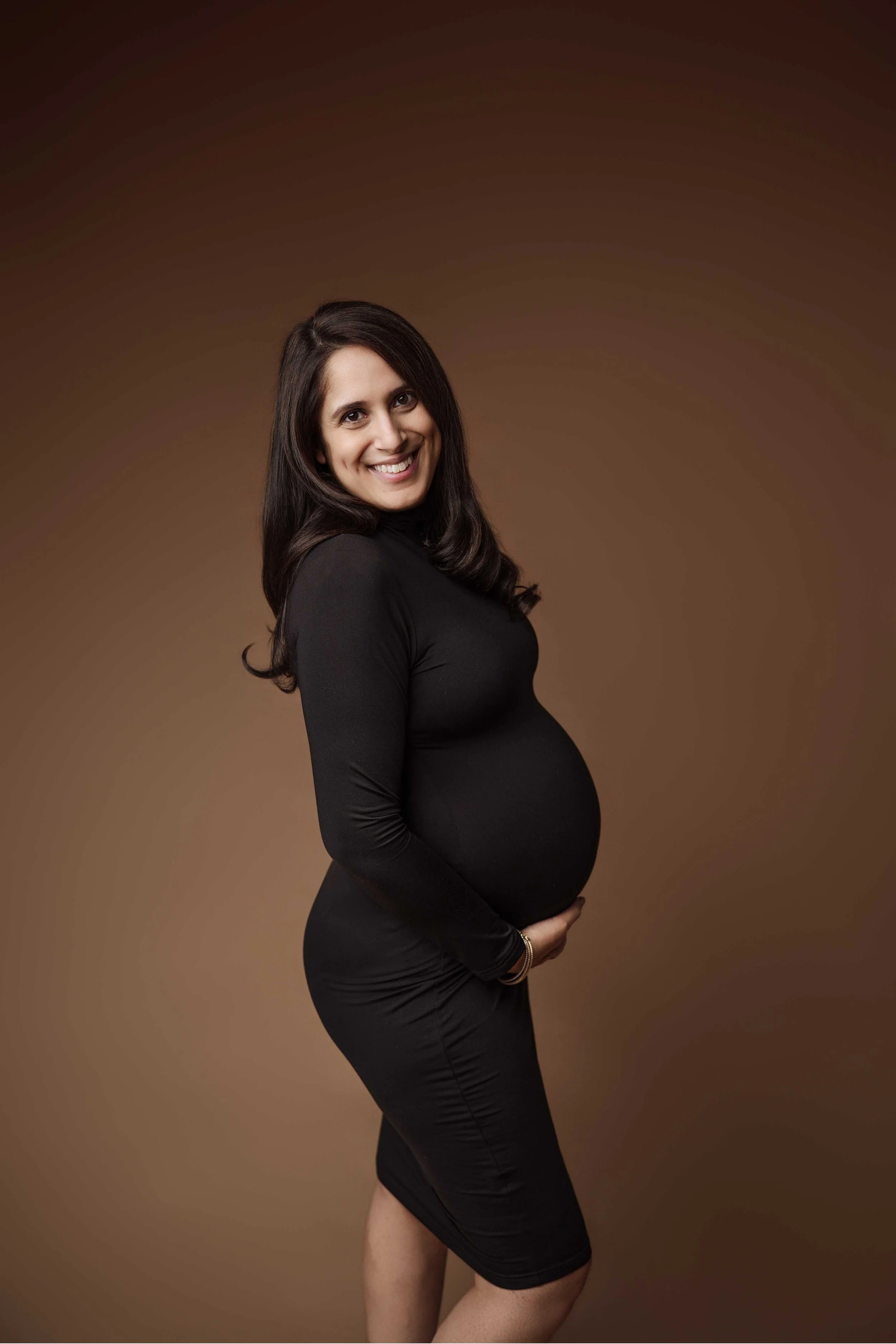 pregnant woman hugs bump and poses for camera, wears black dress