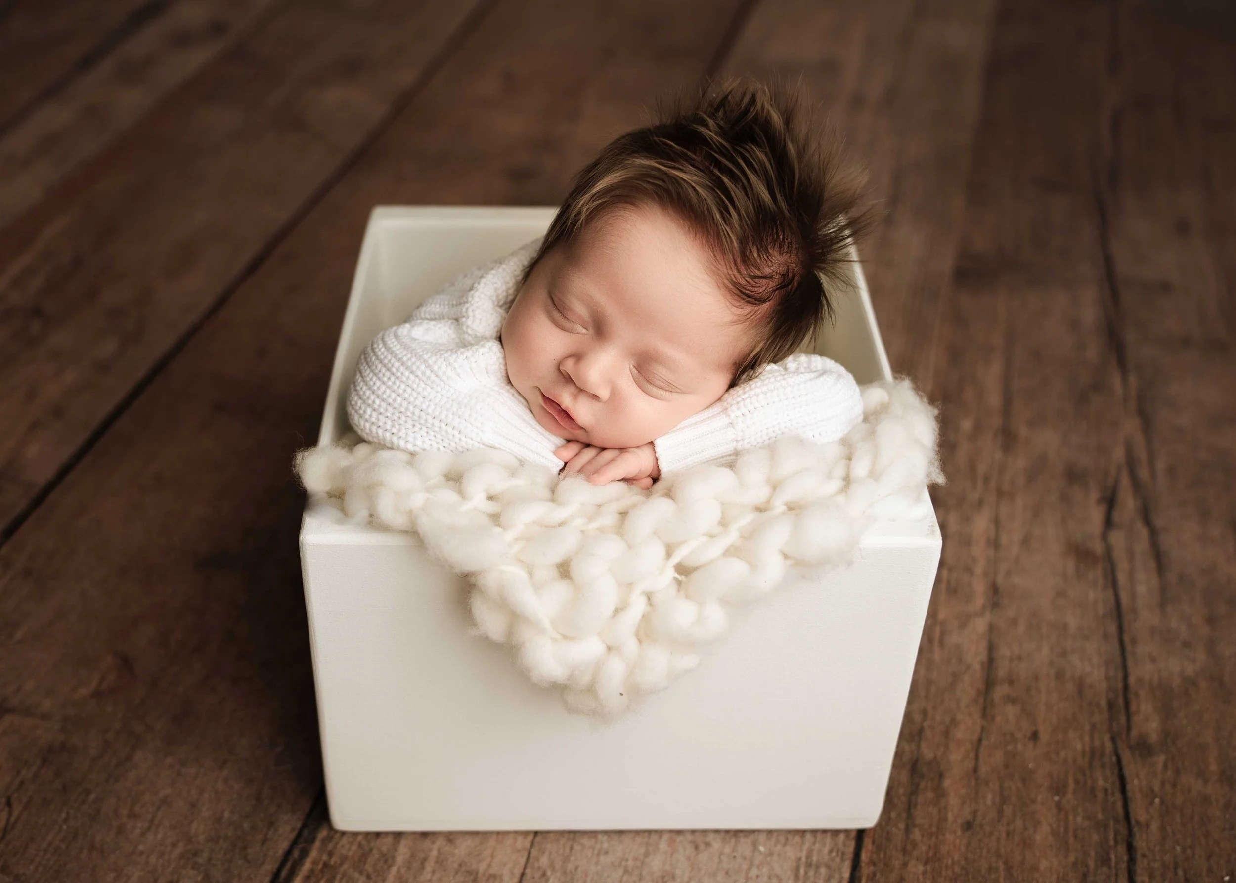 baby in white sweater sleeps in a cream box against a wooden floor