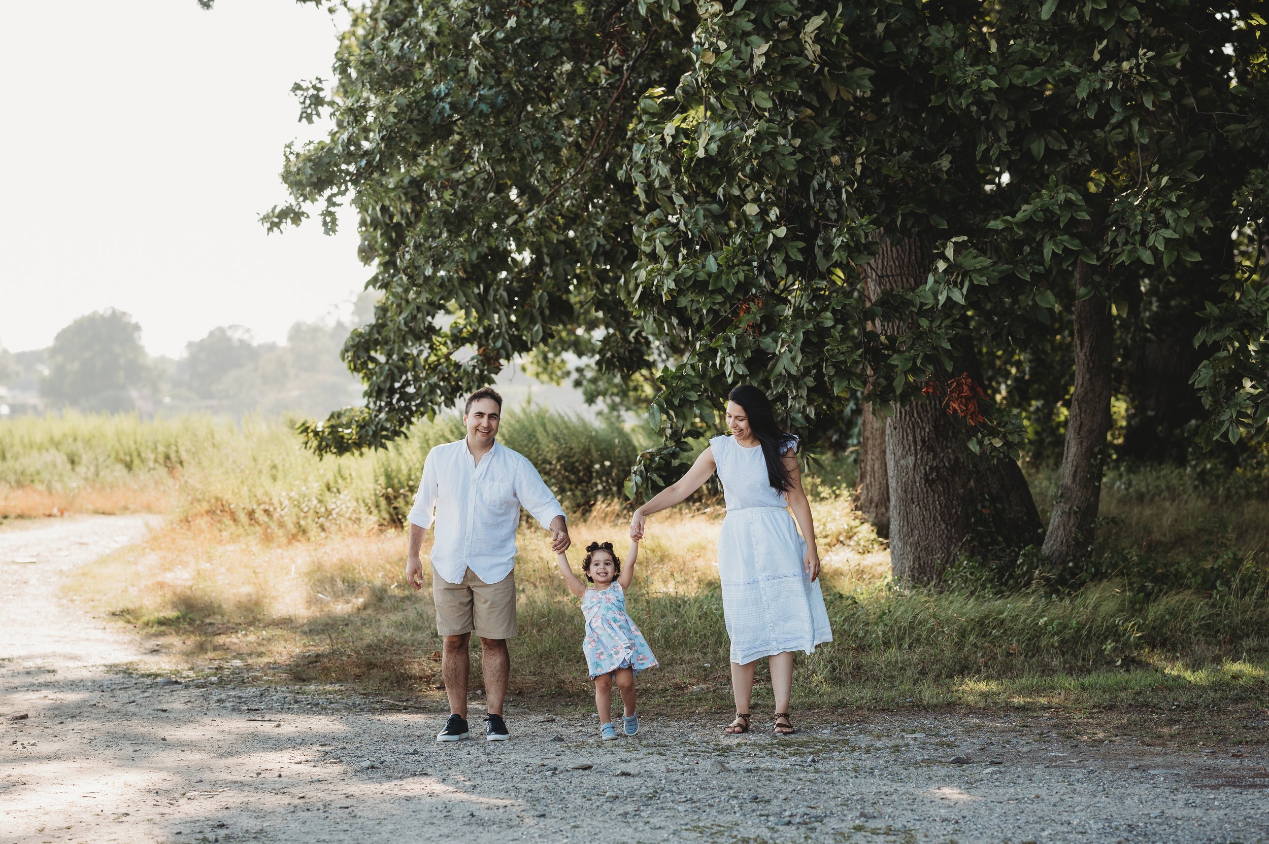 Mom and dad wearing neutral colors hold young daughter's hands on each side in a setting with trees and beach