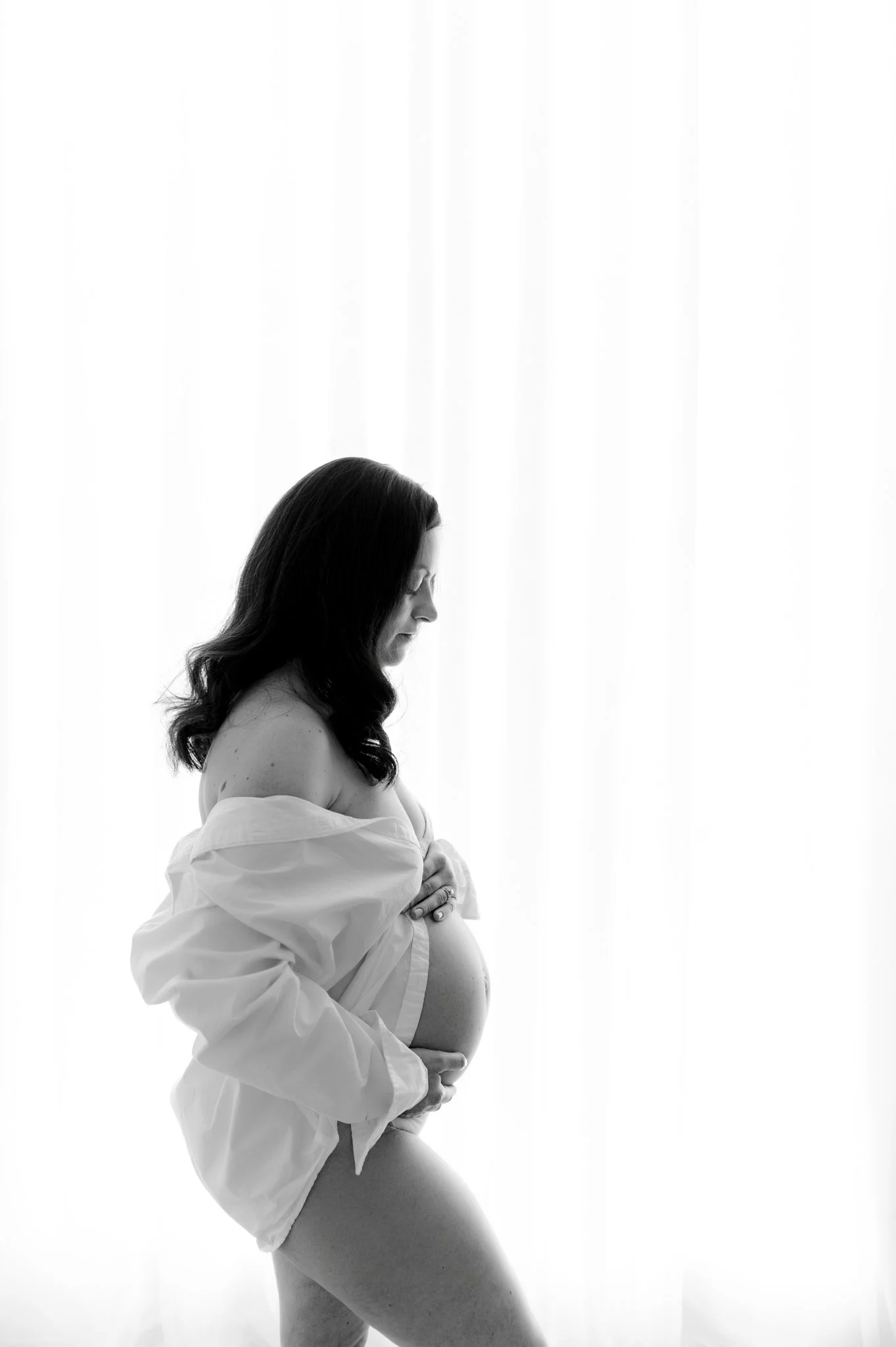 woman in profile cradles pregnant belly wearing oversized white button down