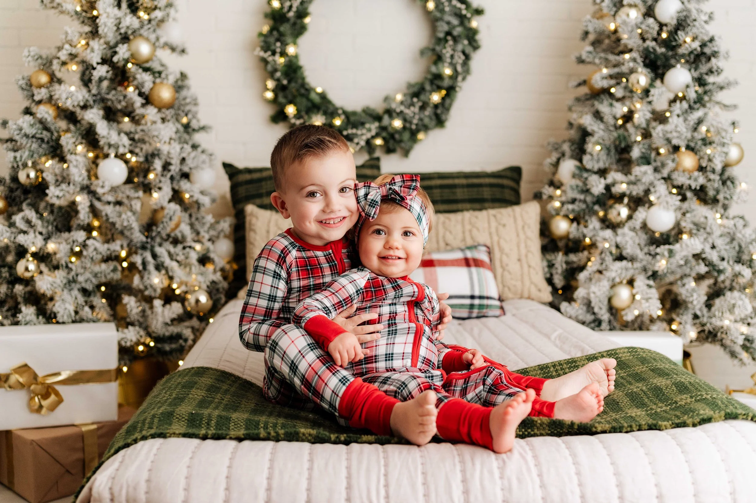siblings in matching plaid pajamas sit on a christmas styled bed scene