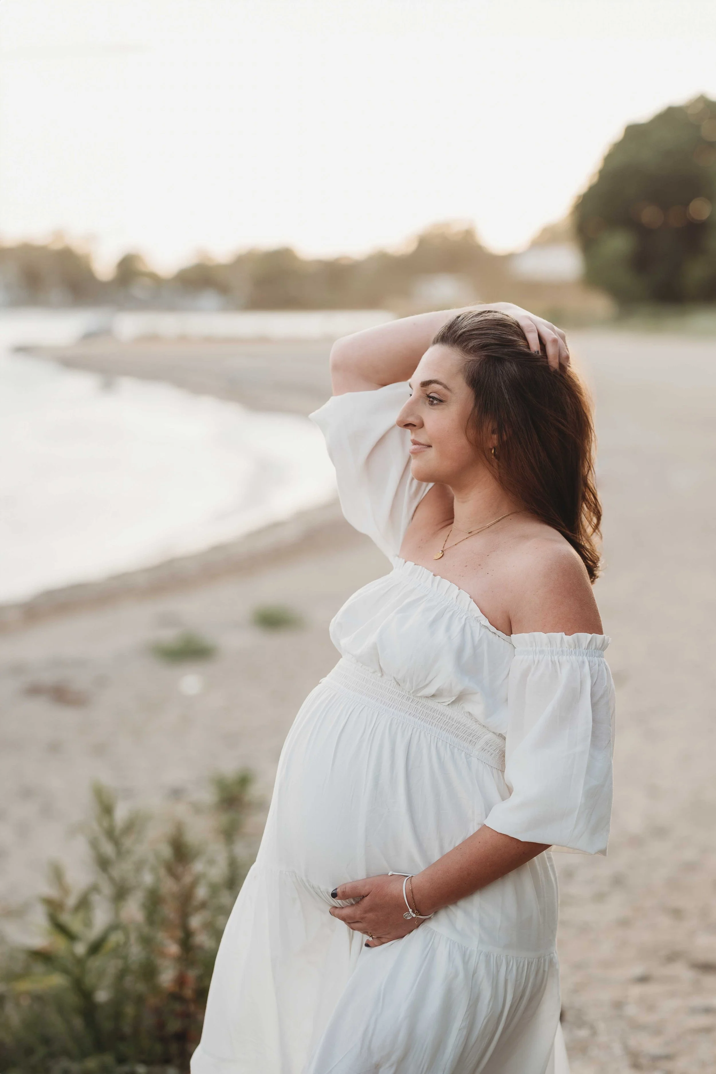 pregnant woman holds hair back and gazes at beach and Long Island sound, Westport maternity photoshoot