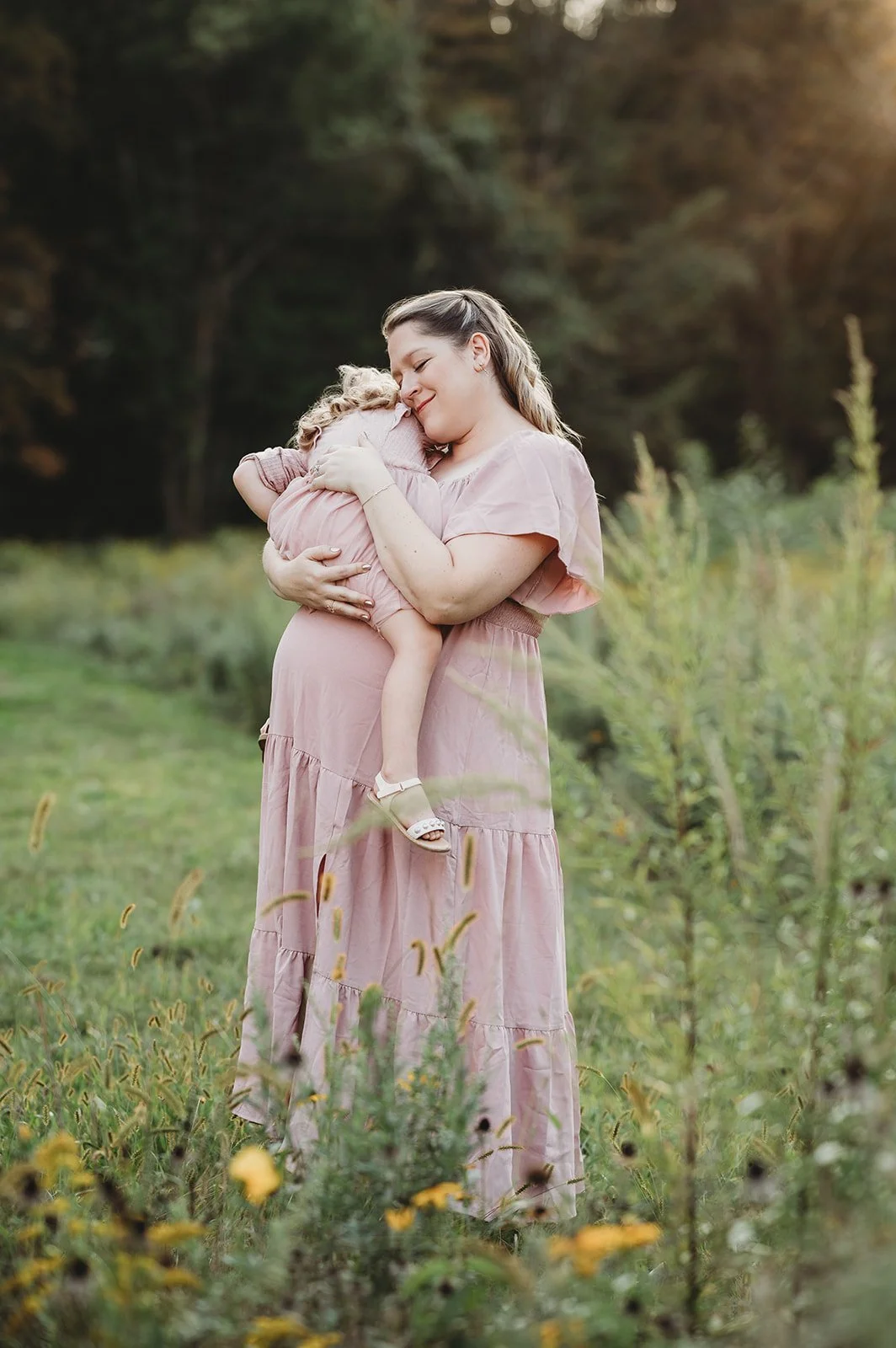 pregnant woman in pink dress snuggles toddler daughter in field of flowers, wilton ct