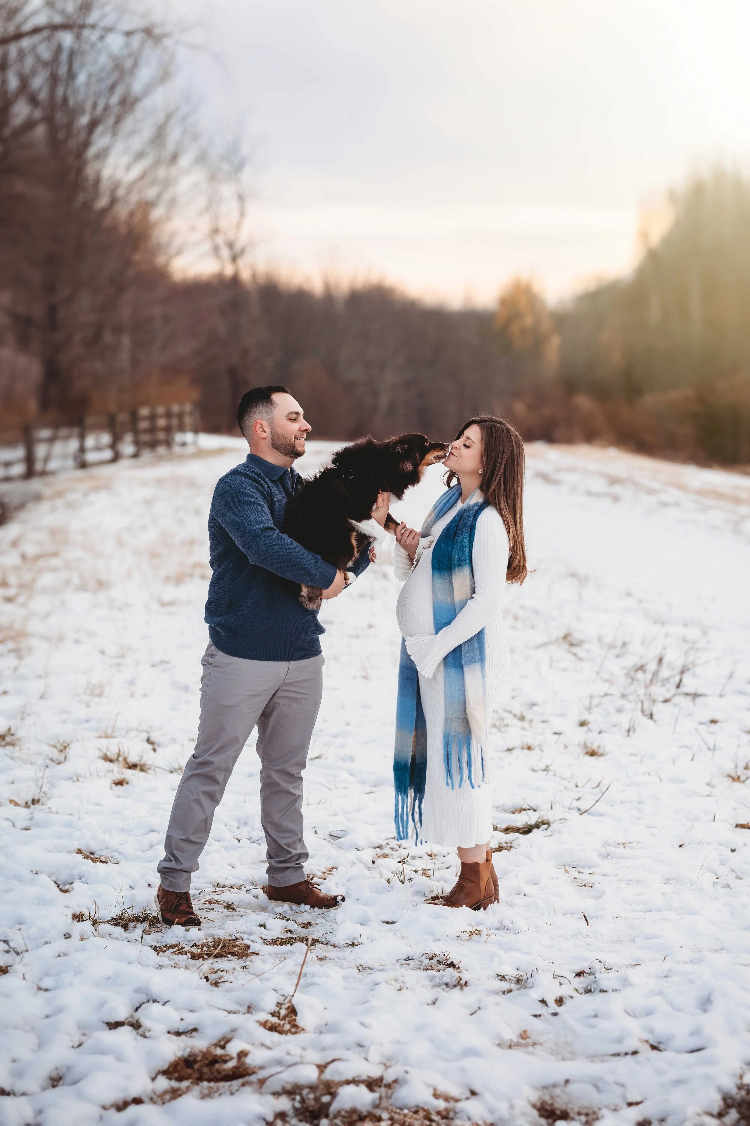 pregnant woman gets kissed by dog, held by husband, in snowy field, golden hour