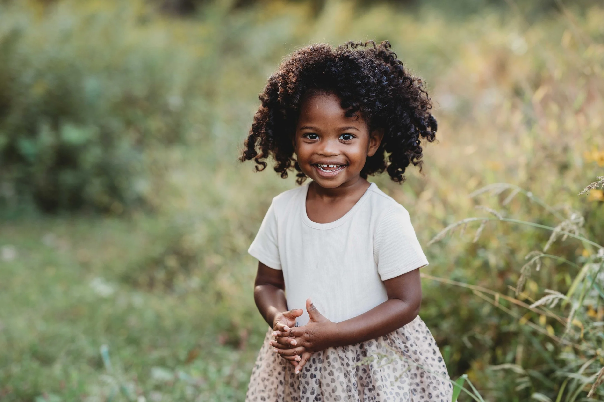 little girl stands in a wilton, ct field wearing a dress and smiling at the camera