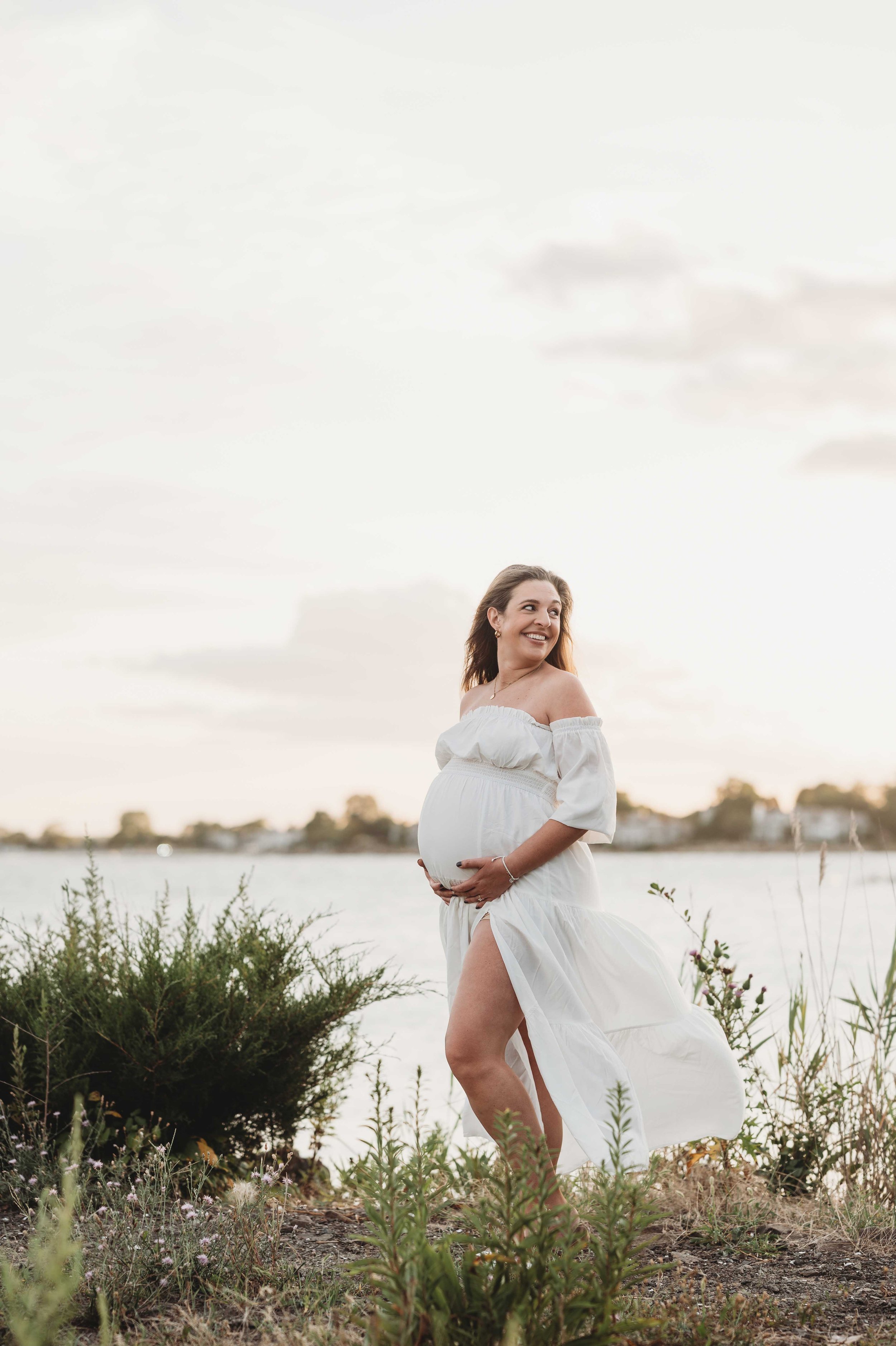Woman wearing flowing white maternity dress poses for maternity photoshoot, westchester, ny