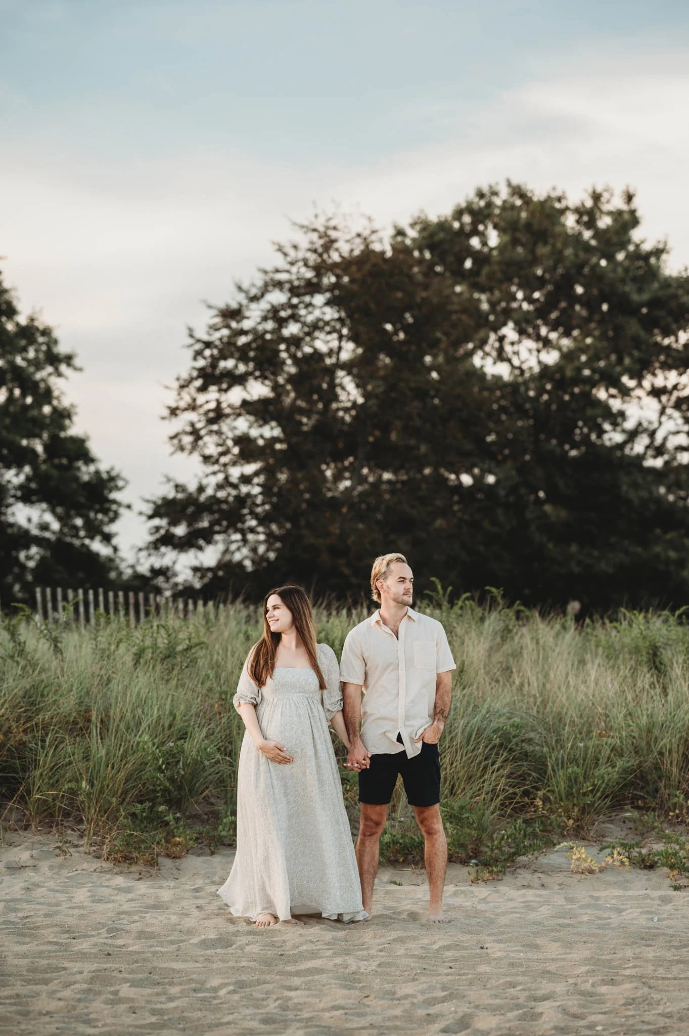 pregnant woman and husband pose in front of beach grass for maternity photoshoot