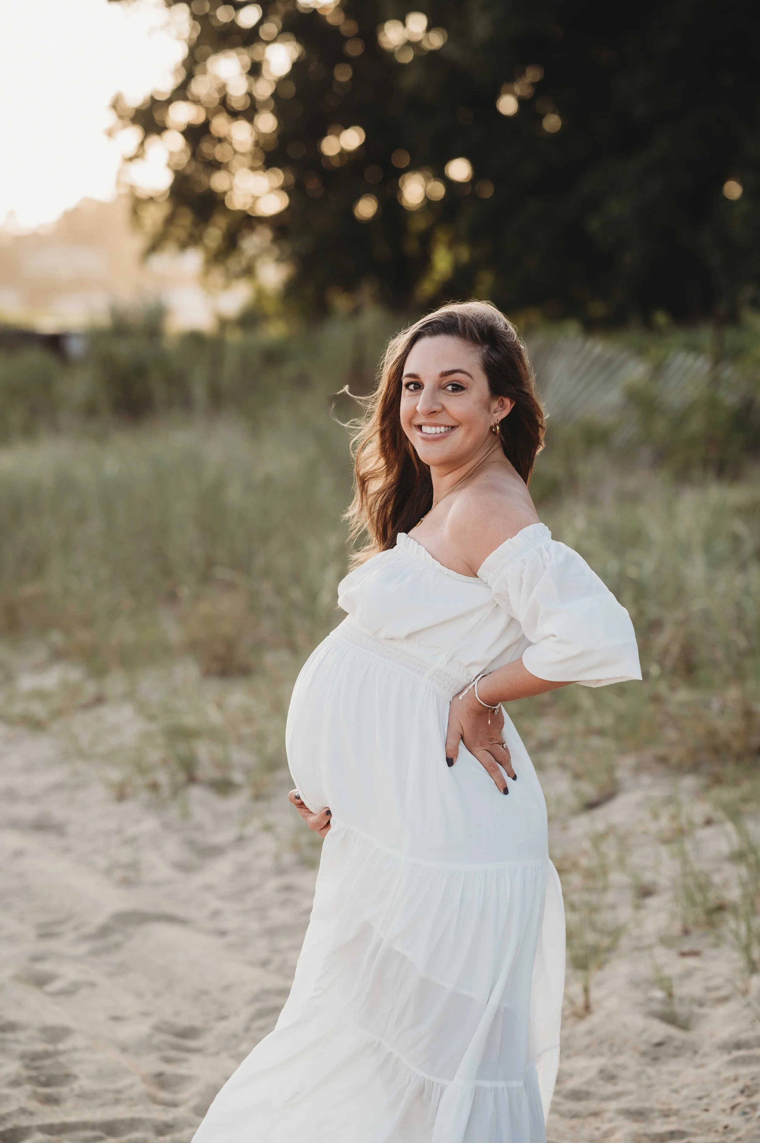 pregnant woman in white dress poses on a Fairfield county, ct beach for a maternity photoshoot