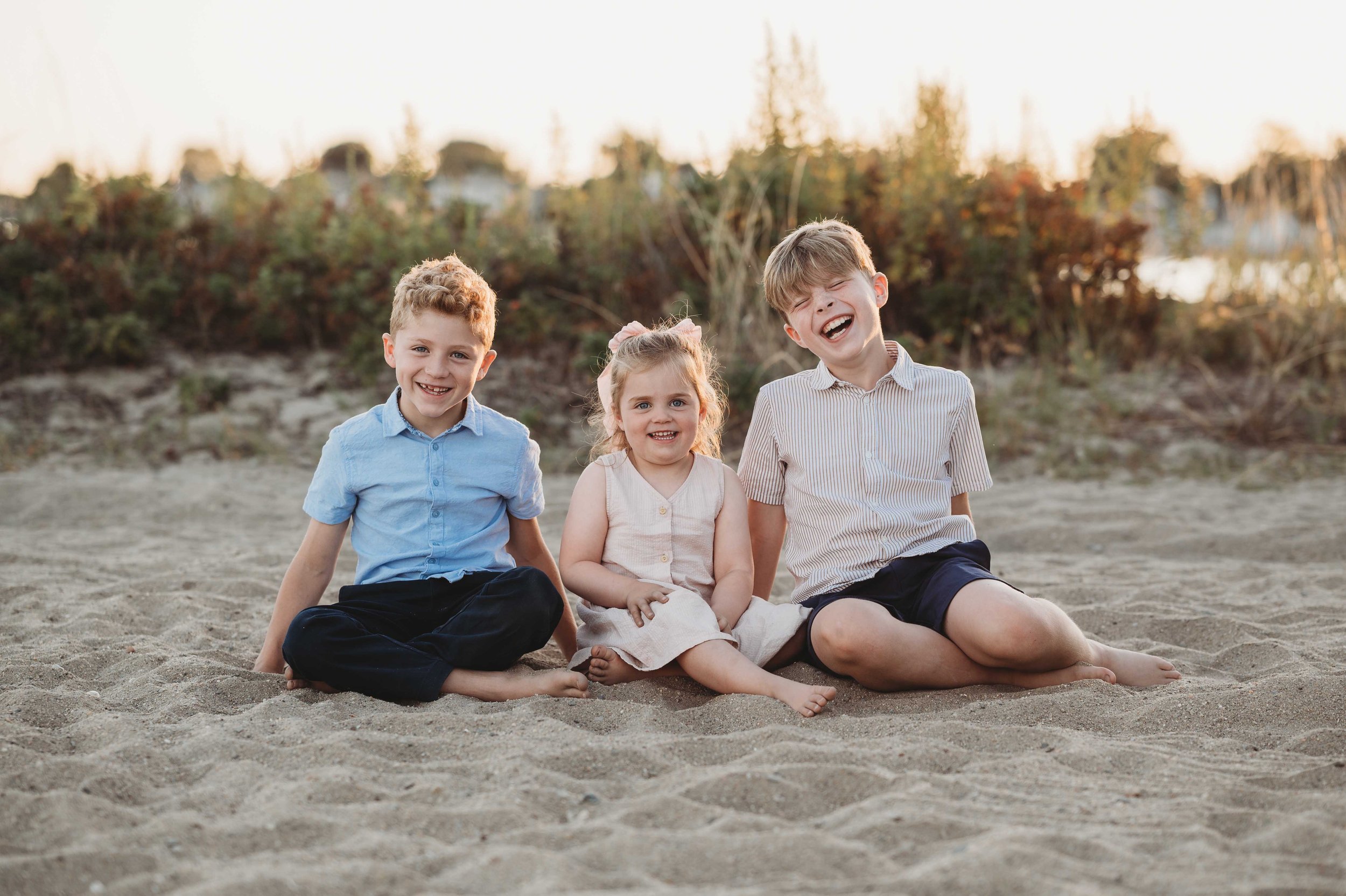three siblings smile and laugh while sitting on a beach in Westport, ct