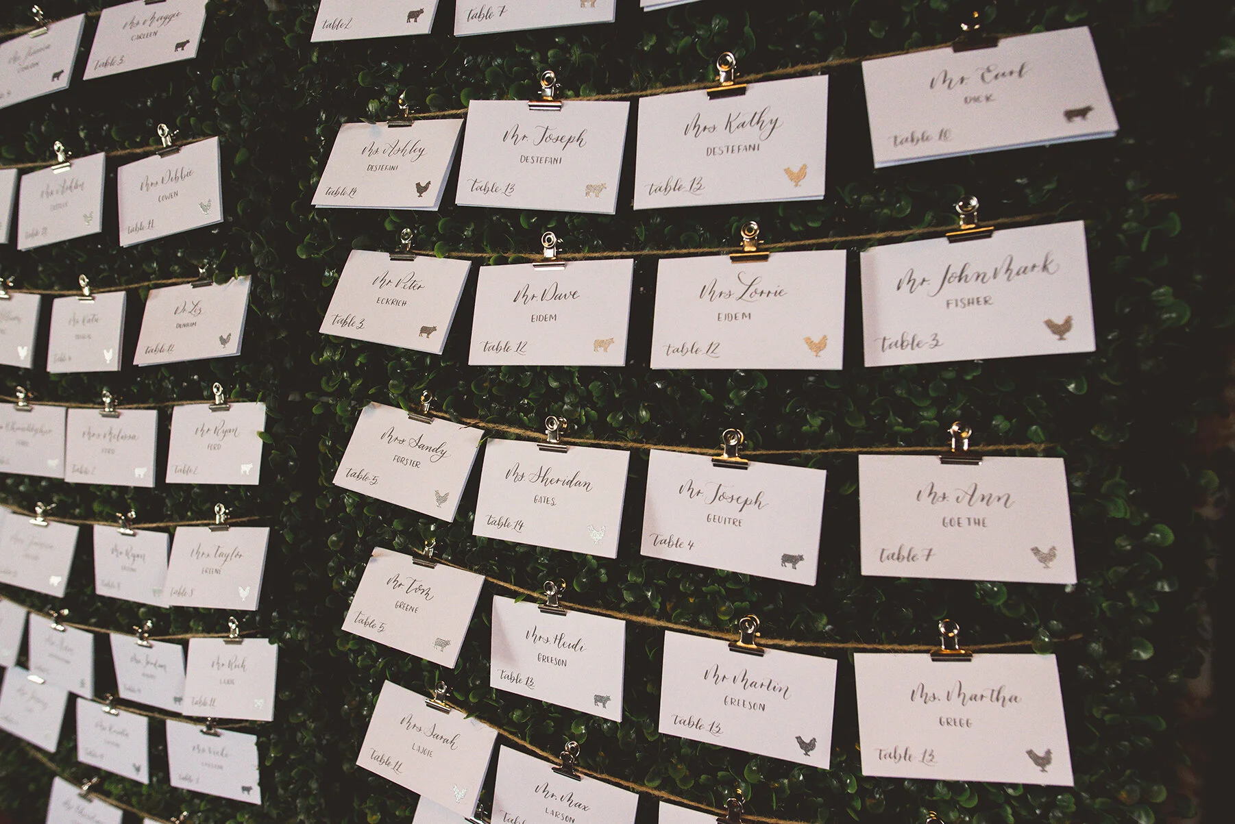 Escort Card display with table numbers and meal selections on a hedge wall.