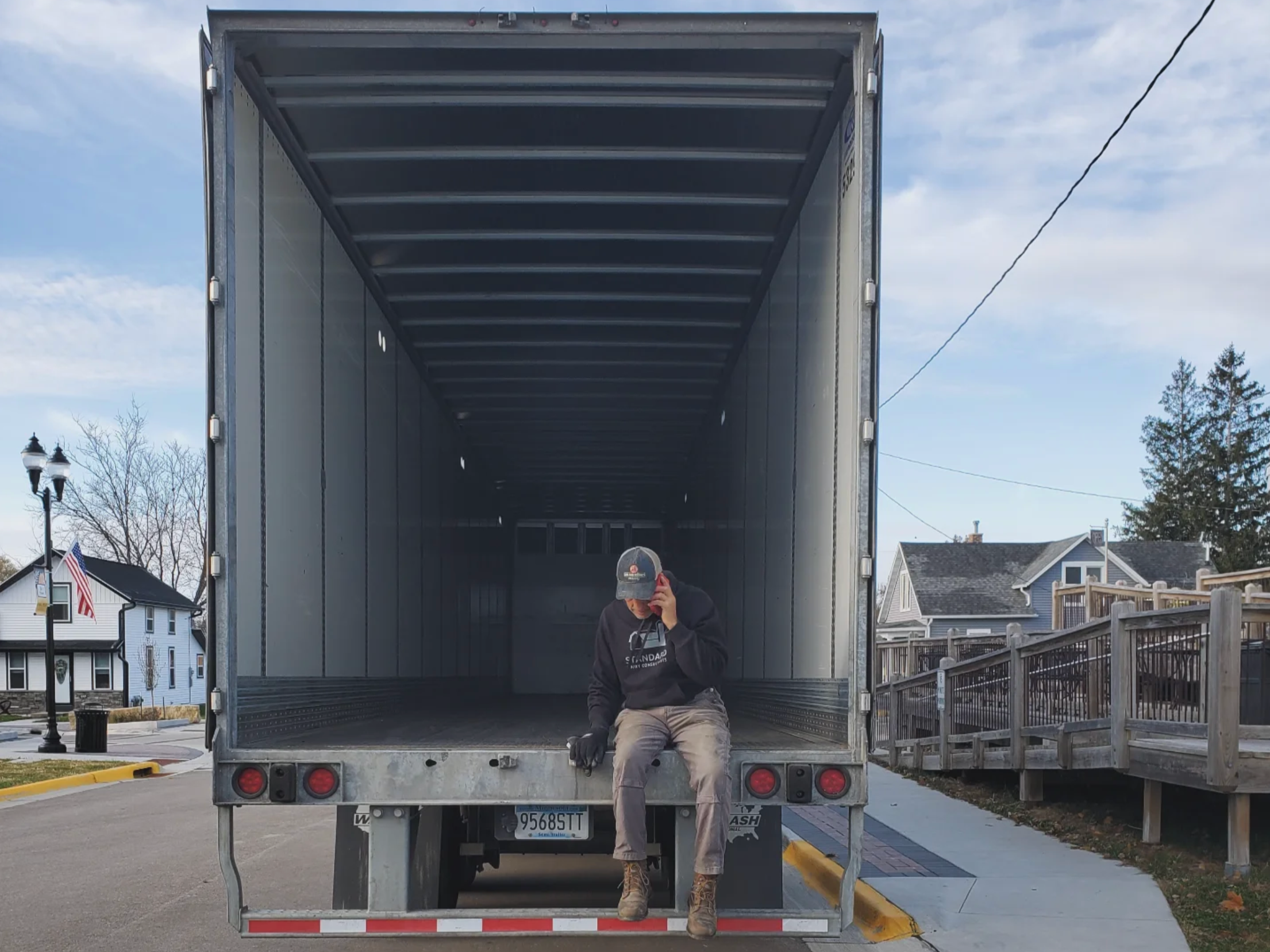 A farmer sits on the back of an empty semi-trailer on a residential street, managing farm deliveries on his own via a phone call.