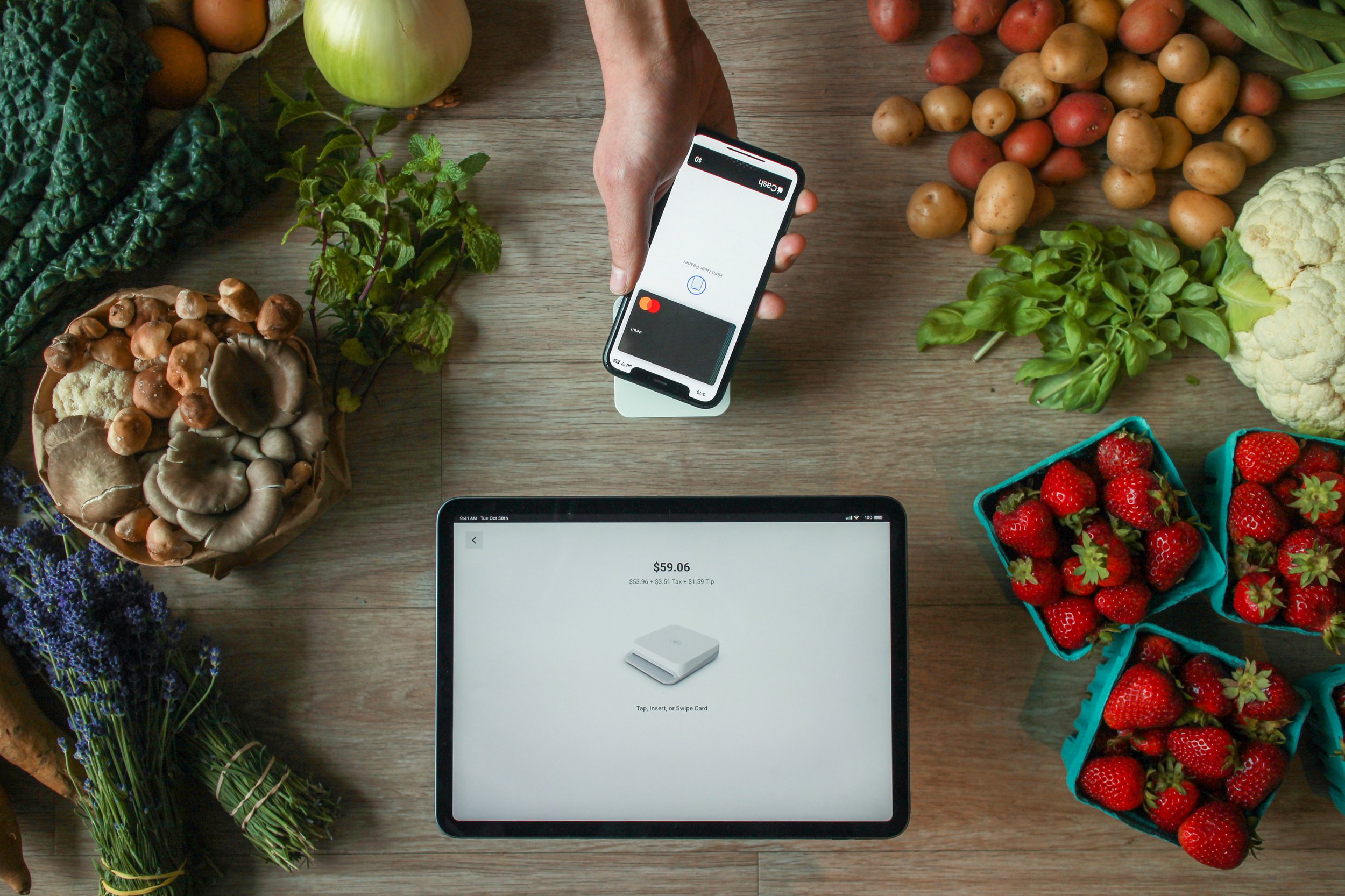 An overhead view of a person using a smartphone to make a contactless payment on a small card reader, next to a tablet displaying a checkout total of $59.06, all surrounded by fresh market produce like strawberries, mushrooms, and potatoes.