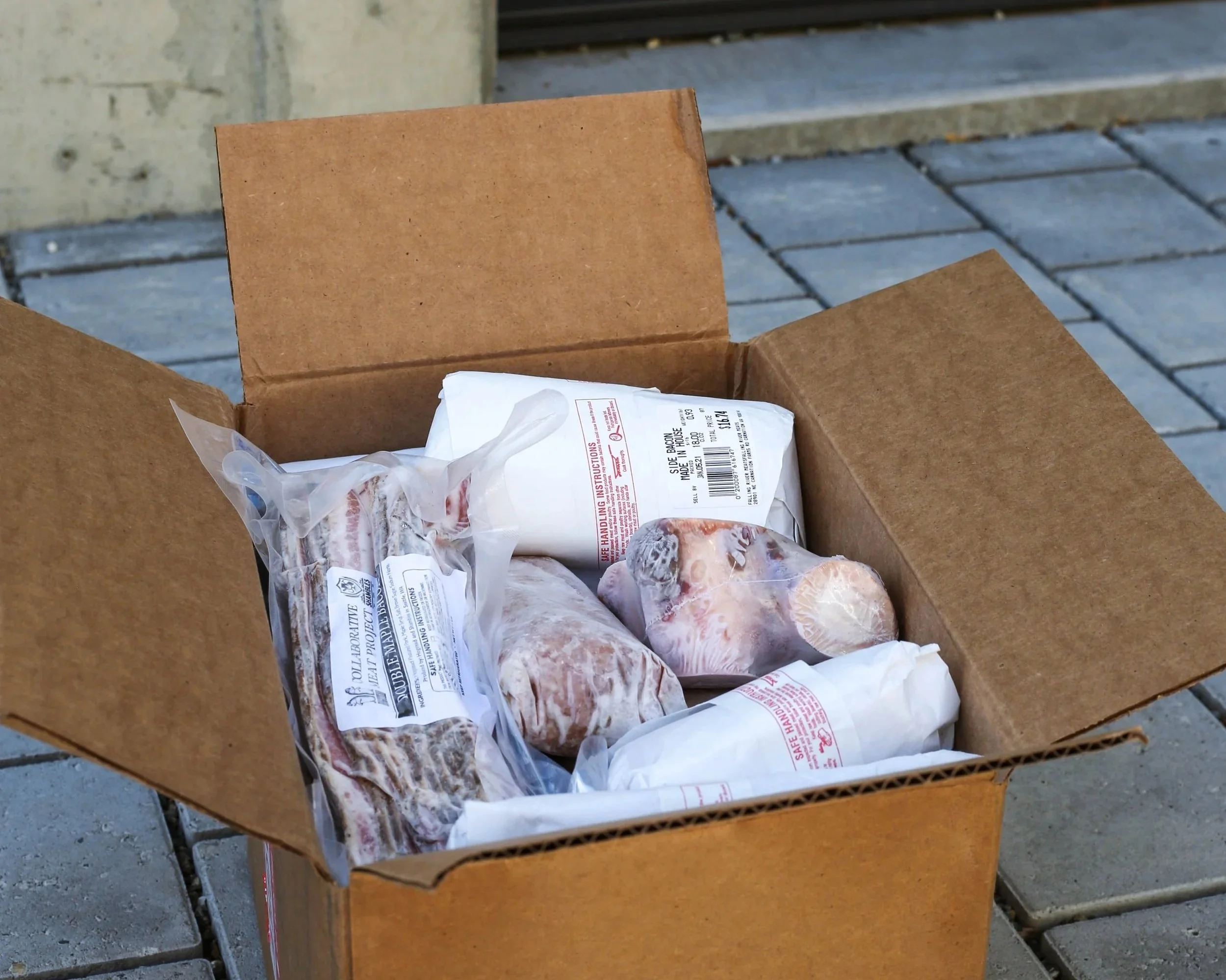 An open cardboard delivery box filled with vacuum-sealed and paper-wrapped packages of meat sitting on a paved patio.