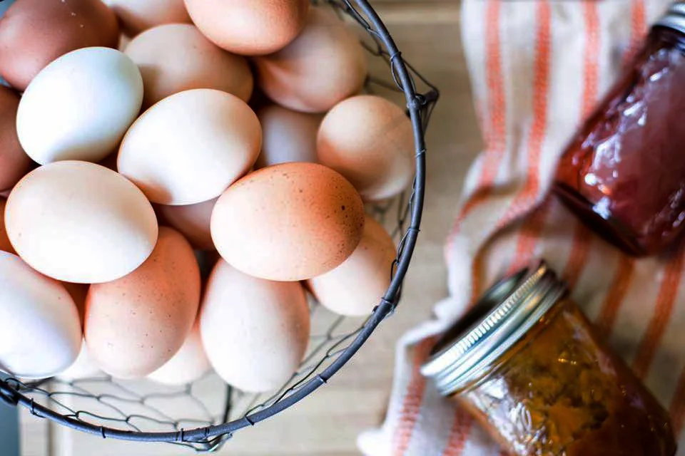 A wire basket filled with fresh, multicolored eggs next to glass jars and a striped kitchen towel.