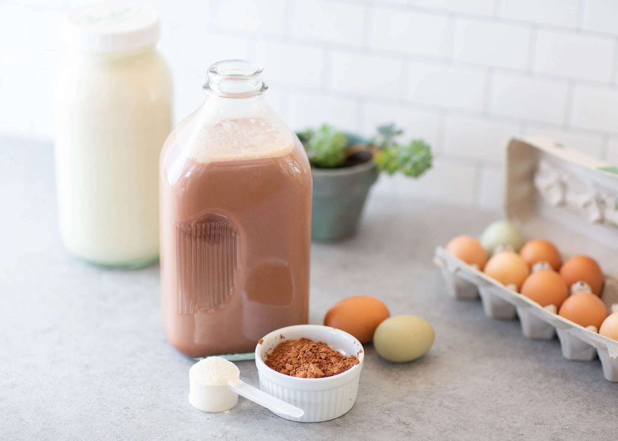 Glass jug of chocolate milk next to a small bowl of cocoa powder, a scoop of powder, and fresh eggs on a kitchen counter.