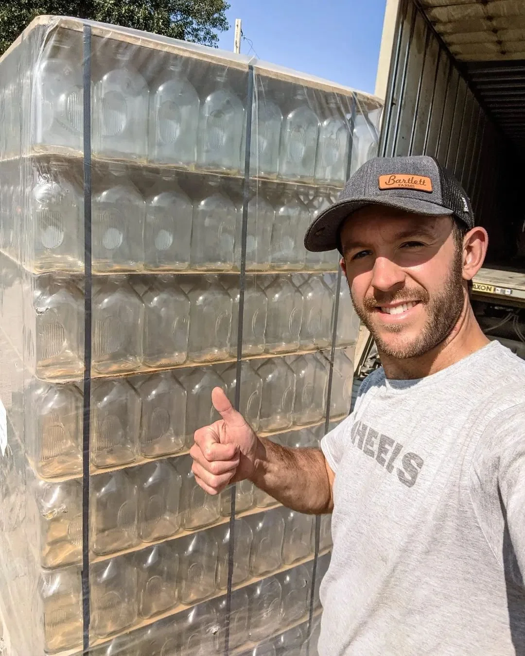 A smiling man in a Bartlett Farms cap giving a thumbs-up next to a large pallet of stacked glass jugs.