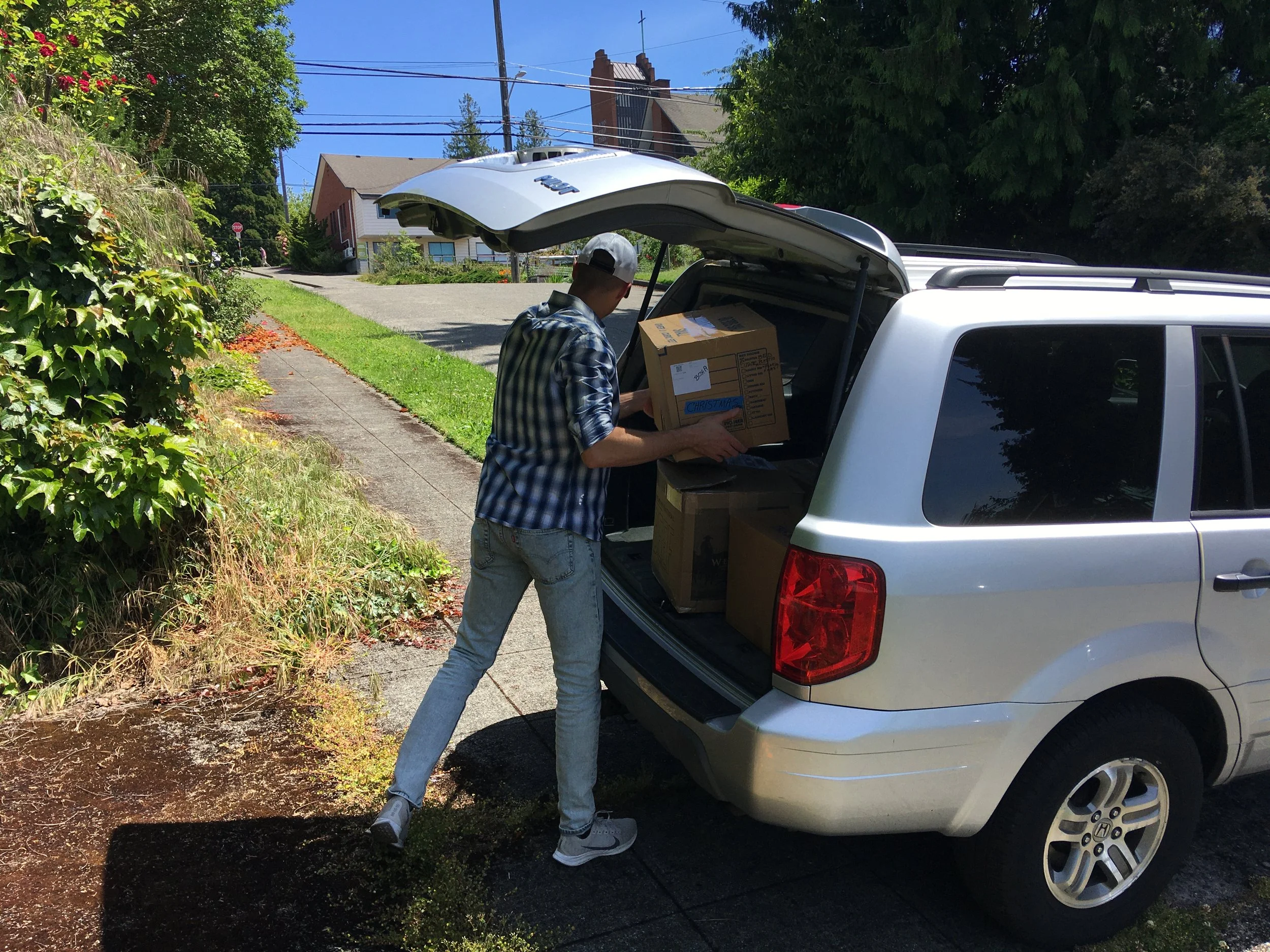 A man loading cardboard boxes into the open trunk of a silver SUV parked on a residential street.