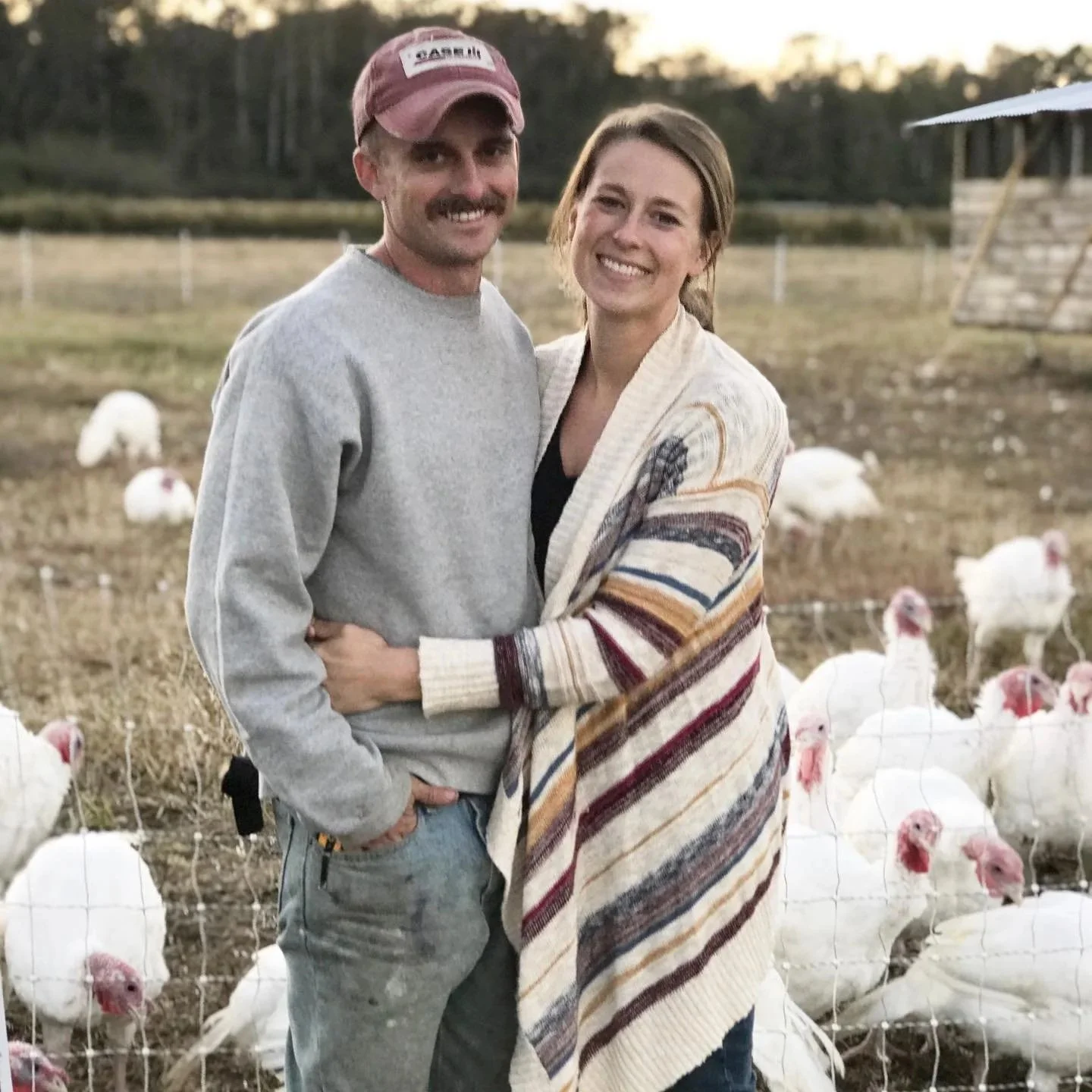 A smiling couple standing together in a pasture surrounded by white turkeys.