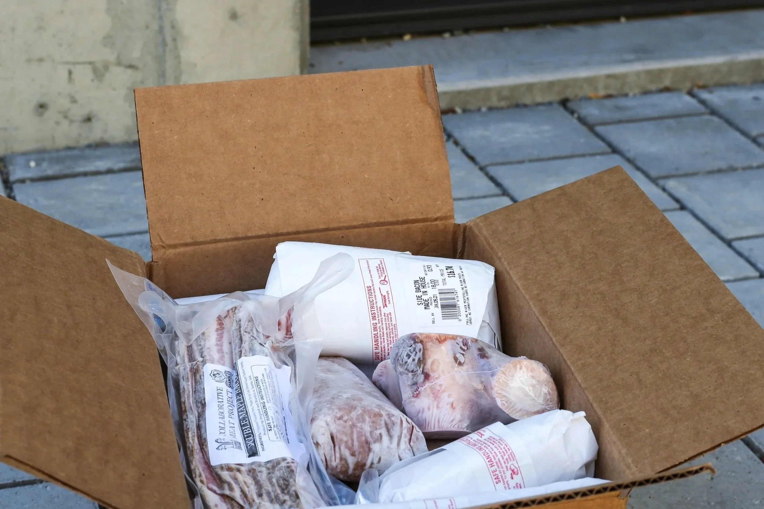 An open cardboard box filled with packages of frozen meat, sitting on a stone patio outside a door.