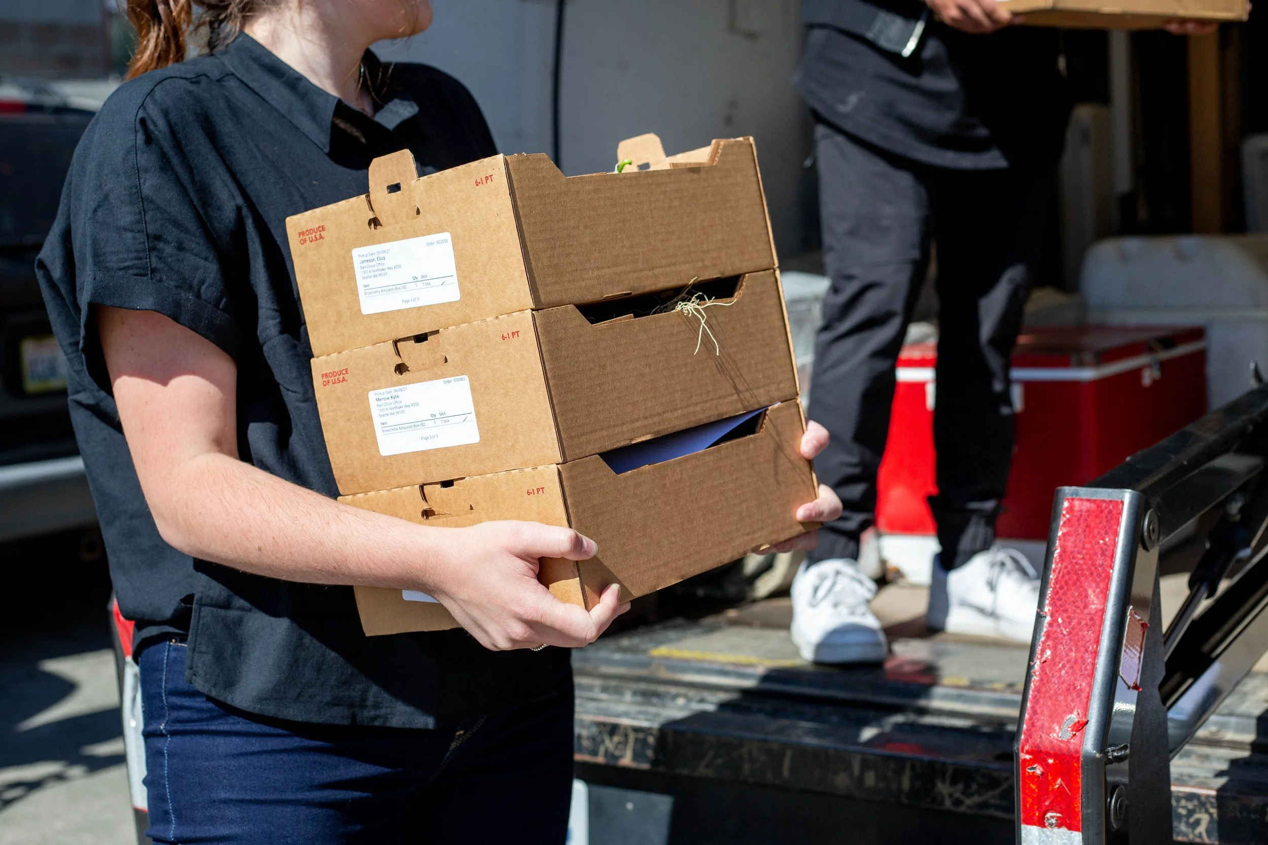 A person carrying a stack of three cardboard produce boxes in front of a delivery truck.
