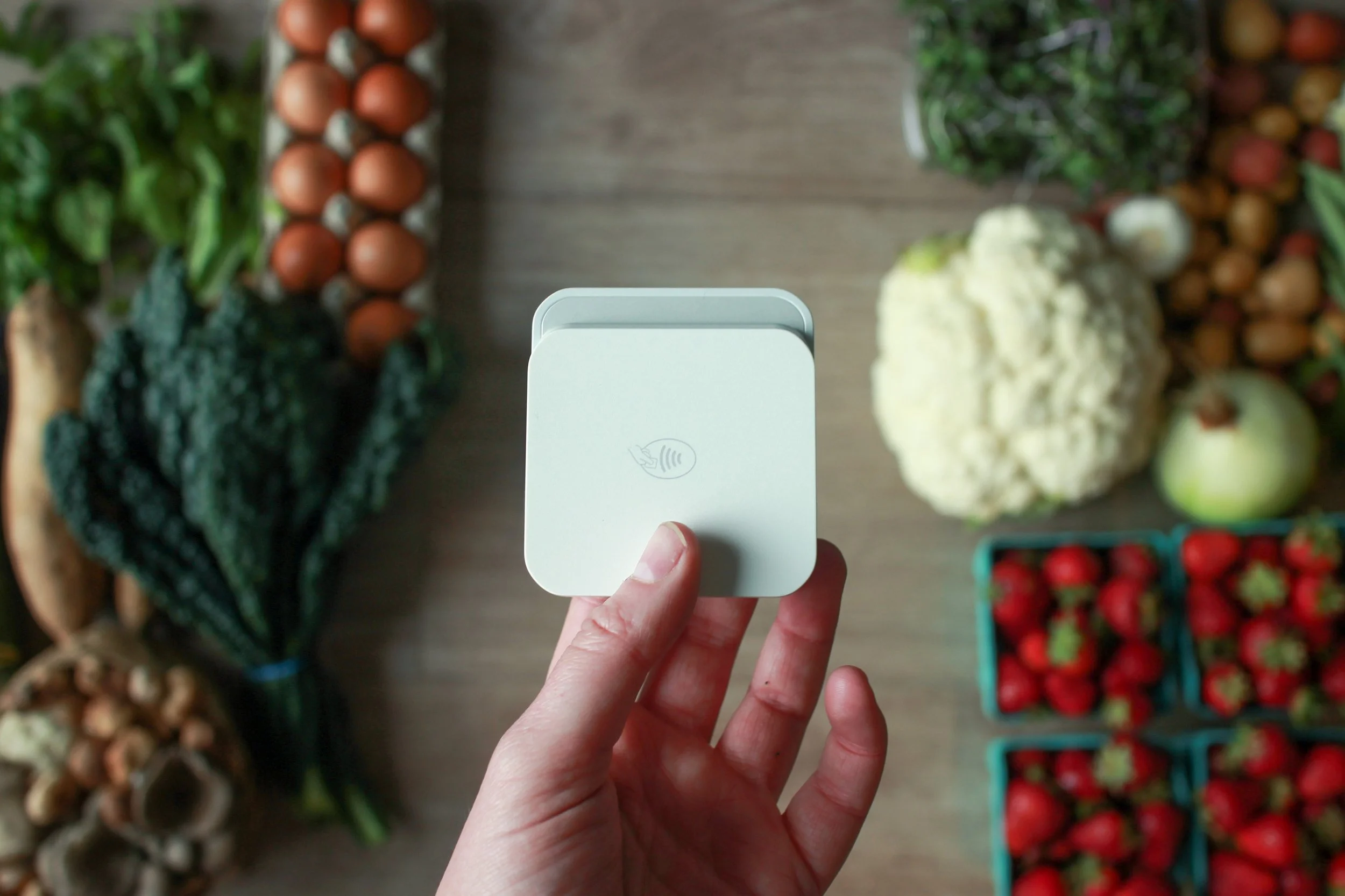 A hand holding a white contactless card reader over an assortment of fresh farmers market produce arranged on a wooden surface.