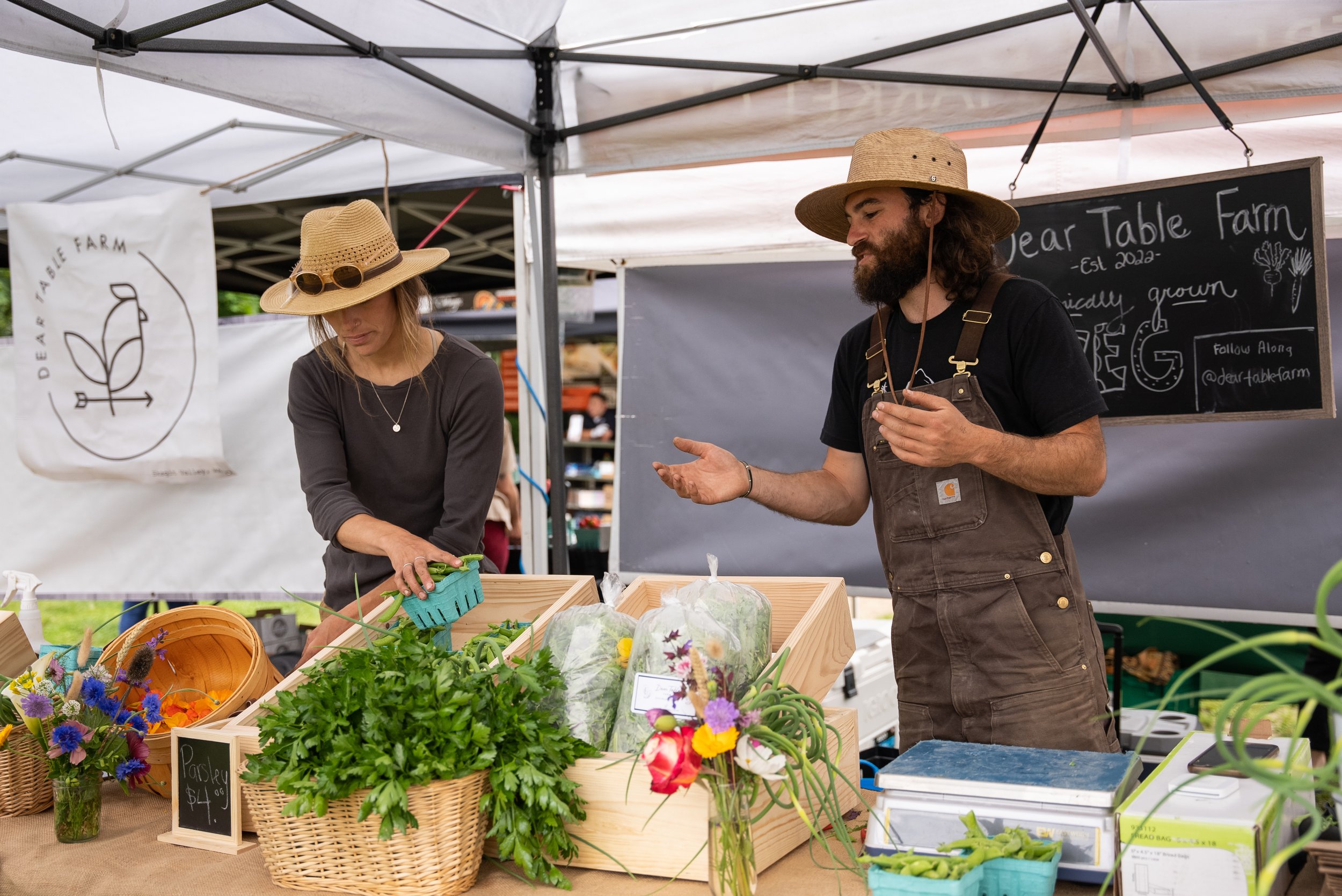 Two farmers wearing straw hats manage a "Dear Table Farm" market stall filled with fresh herbs, produce, and flowers under a white canopy tent.