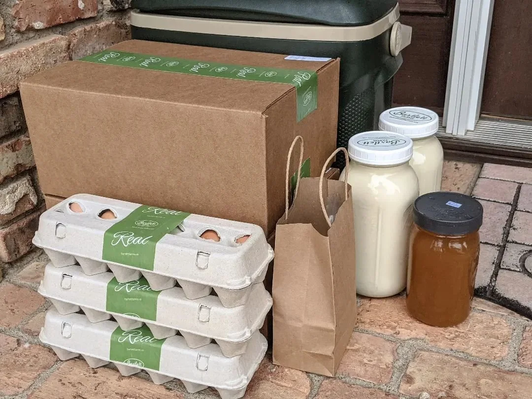 A farm-fresh delivery from a Farm on a brick porch, featuring three stacked cartons of eggs, glass jars of milk and honey, and cardboard packaging.