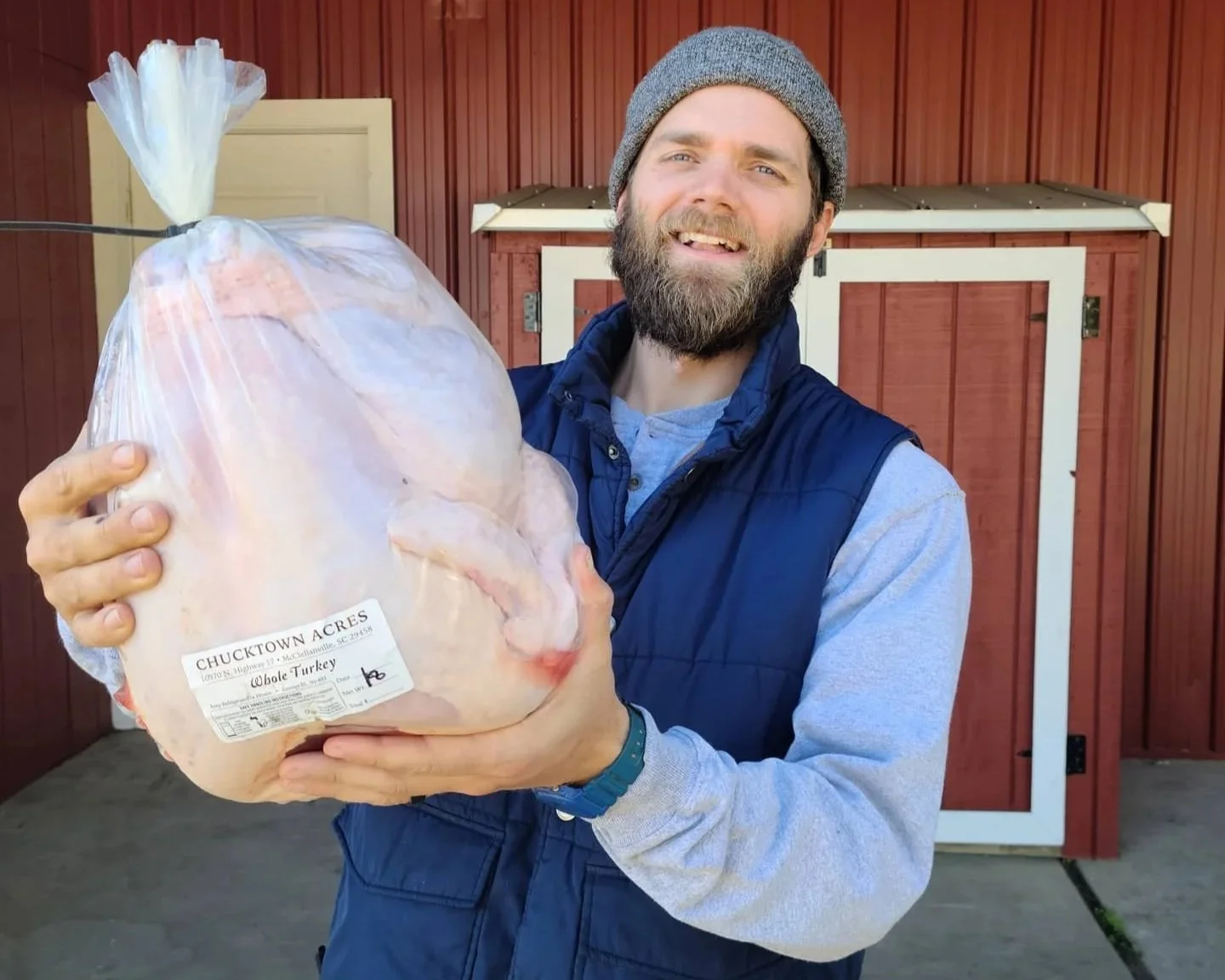 A smiling man holding a large, packaged Chucktown Acres whole turkey in front of a red barn.